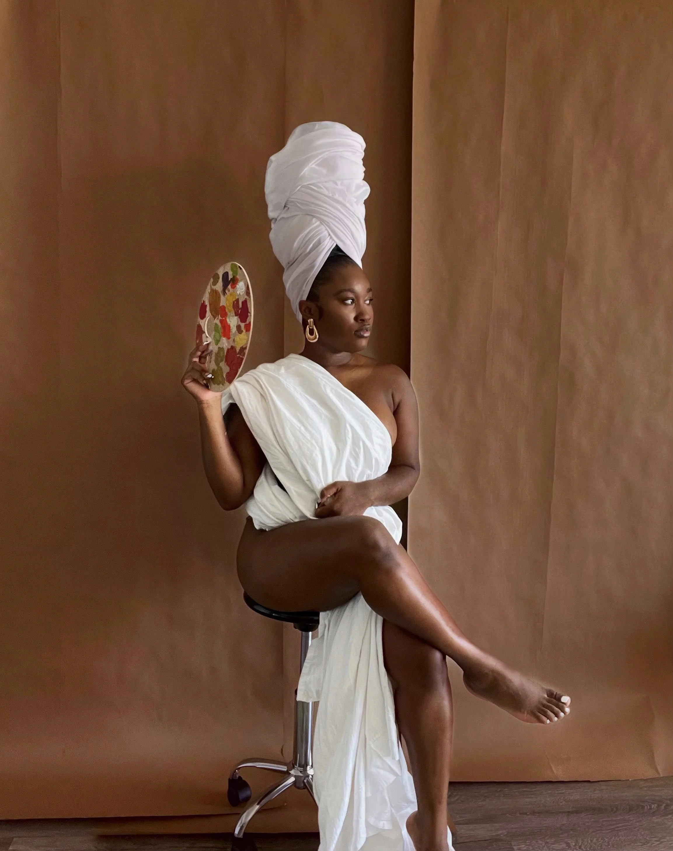 A woman with dark skin wearing a white cloth draped over her body, with a tall white headwrap, sitting on a stool against a brown background. She holds a colorful patterned fan in one hand and looks to the side.