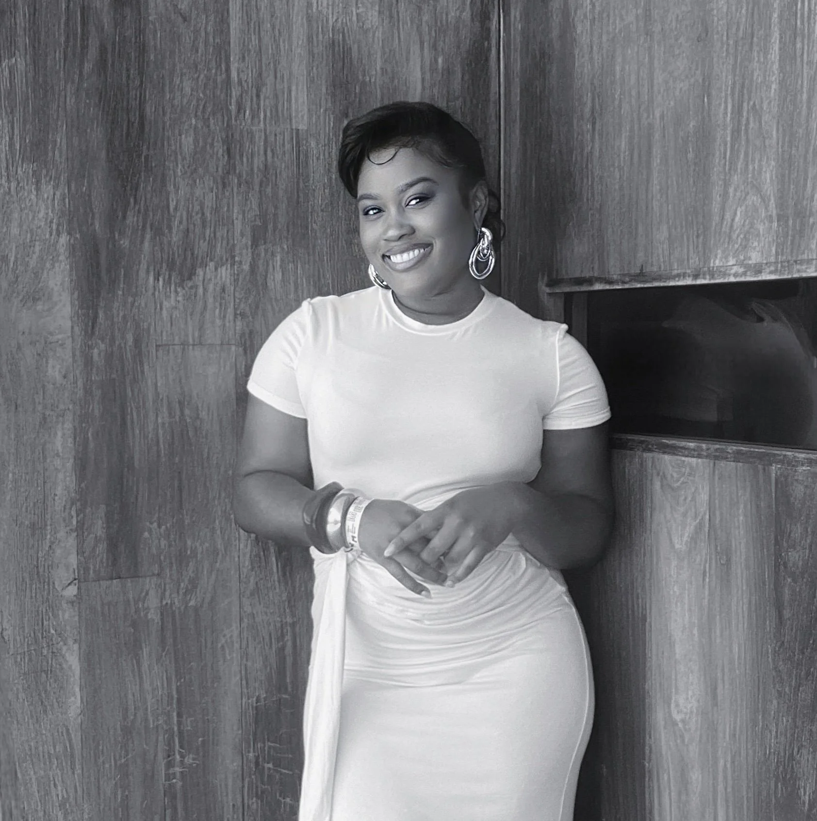 A woman with short dark hair, earrings, and bracelets, smiling and standing against a wooden wall.