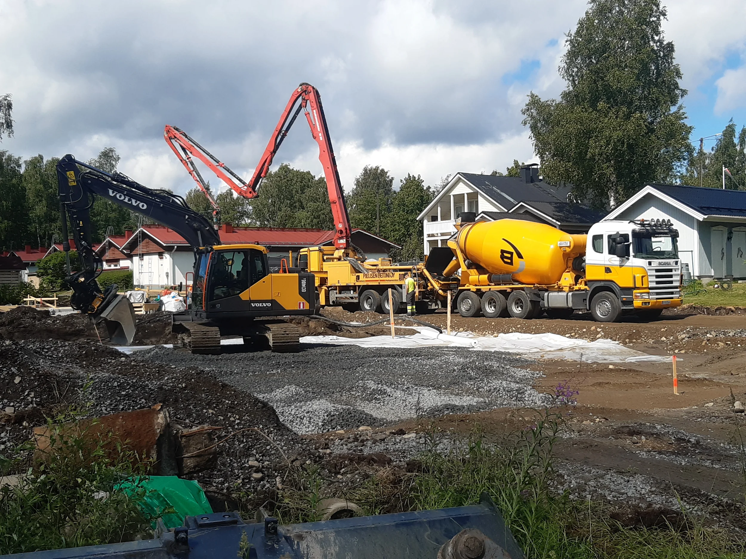 Rakennustyömaa, jossa kaivinkone ja betoninkuljetusauto työskentelevät rakennuksen perustusten parissa. Taustalla taloja ja puita, pilvinen taivas.
