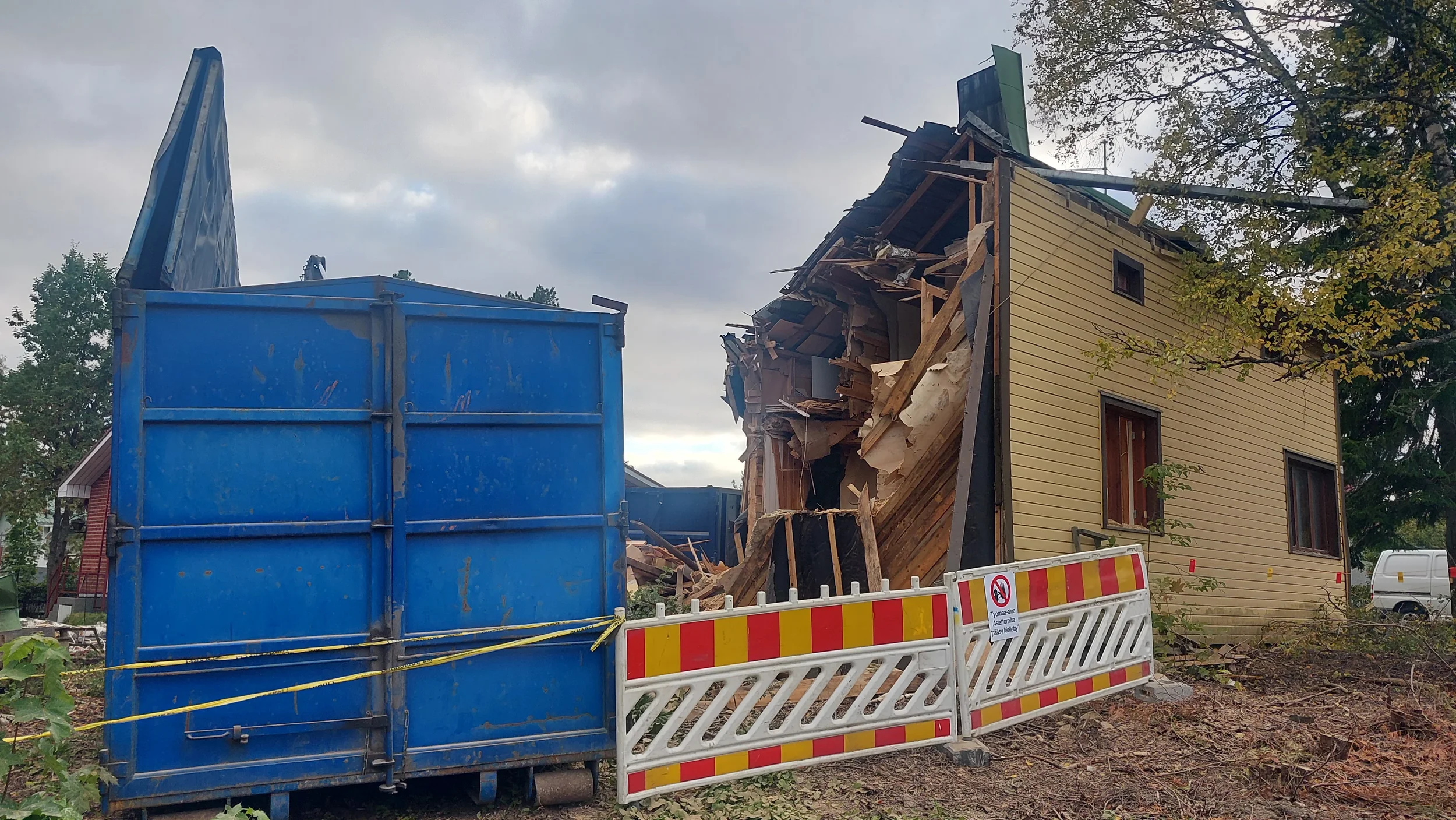 Työmaa-aidan takana puoliksi purettu kaksikerroksinen talo ja roskalava. Taustalla puita ja pilvinen taivas.