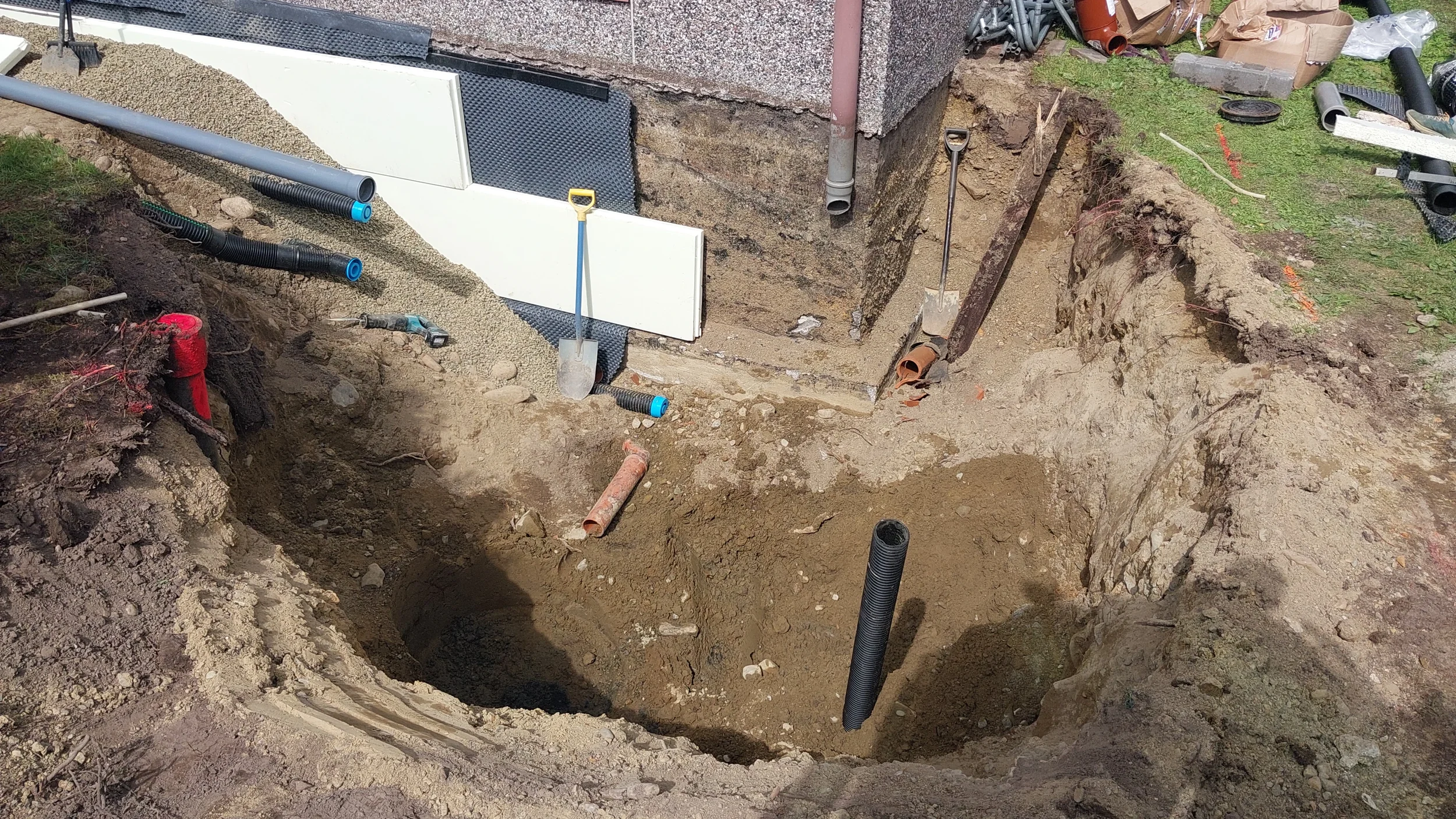 Käynnissä oleva vanhan talon salaojitus, näkyvissä kaivettua maata, putkia, työkaluja ja rakennuksen seinää.