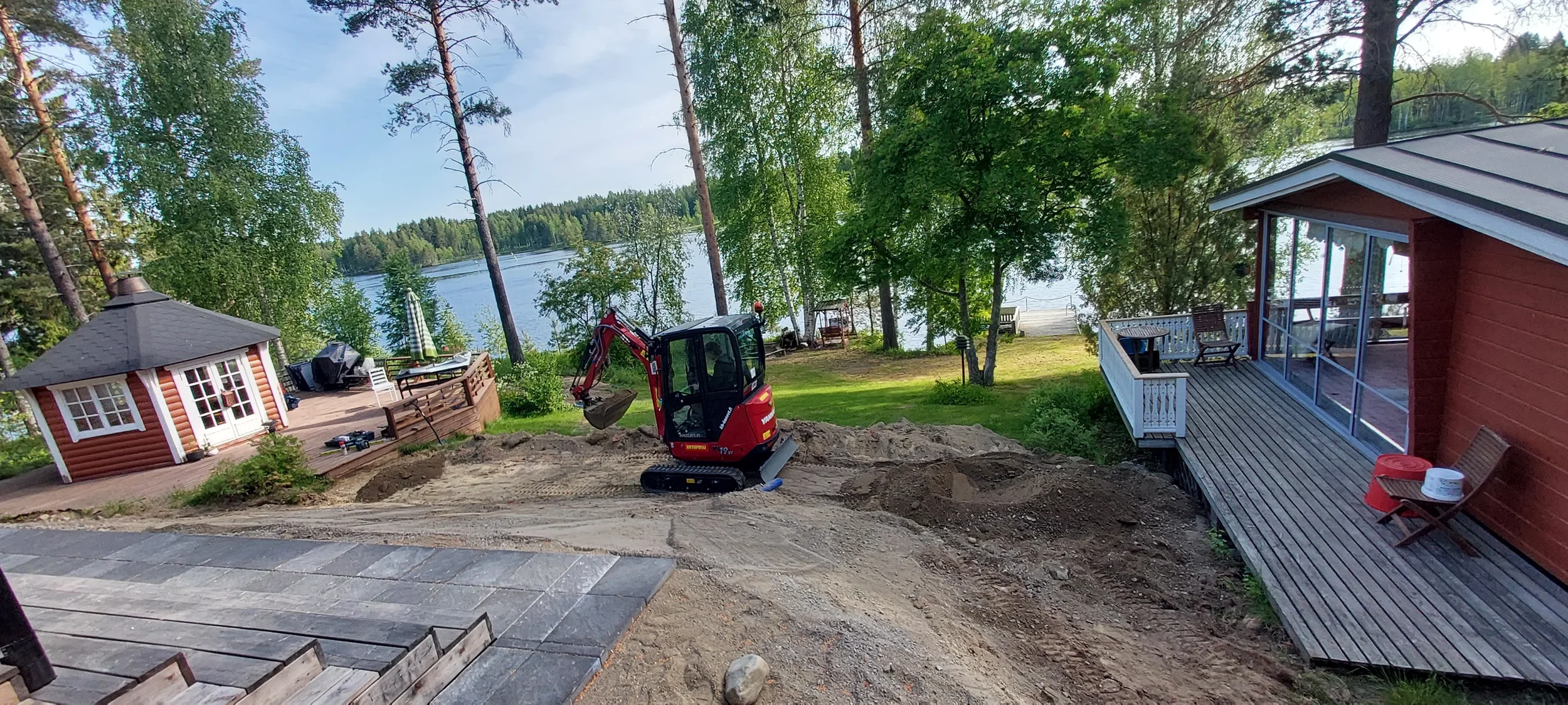 Työn alla oleva mökin piha. Vasemmalla pieni punainen huvimaja, keskellä pieni kaivinkone, oikealla punainen rakennus ja terassi. Ttaustalla pihamaisemaa, järvi ja puita.
