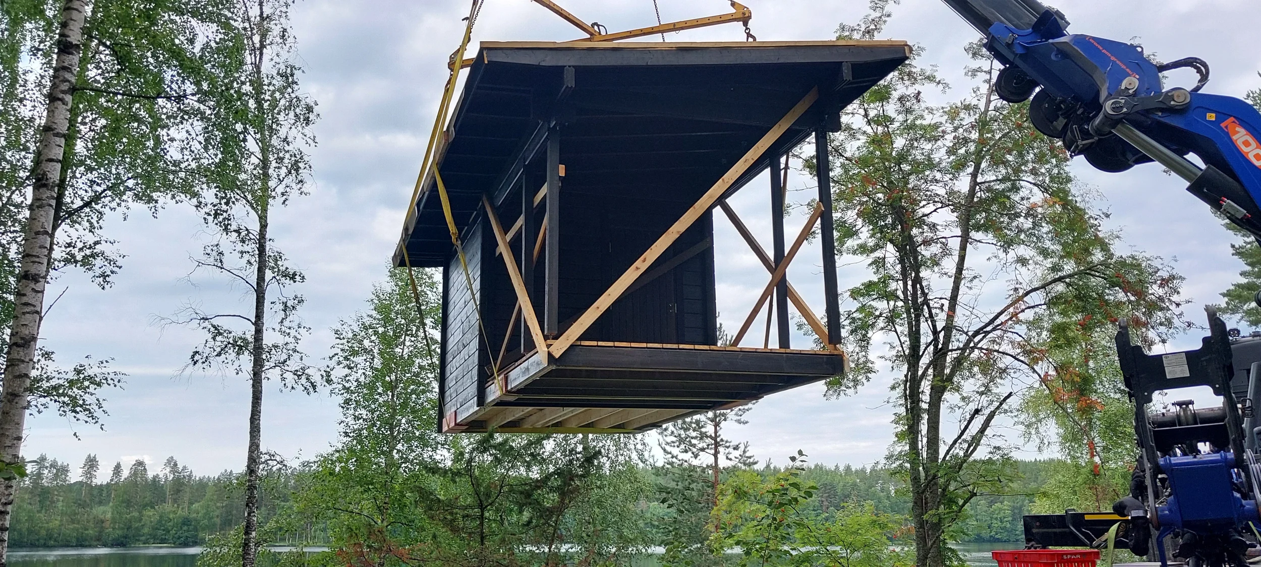 Rantasauna nostetaan nosturilla paikalleen. Taustalla puita ja osittain pilvinen taivas.