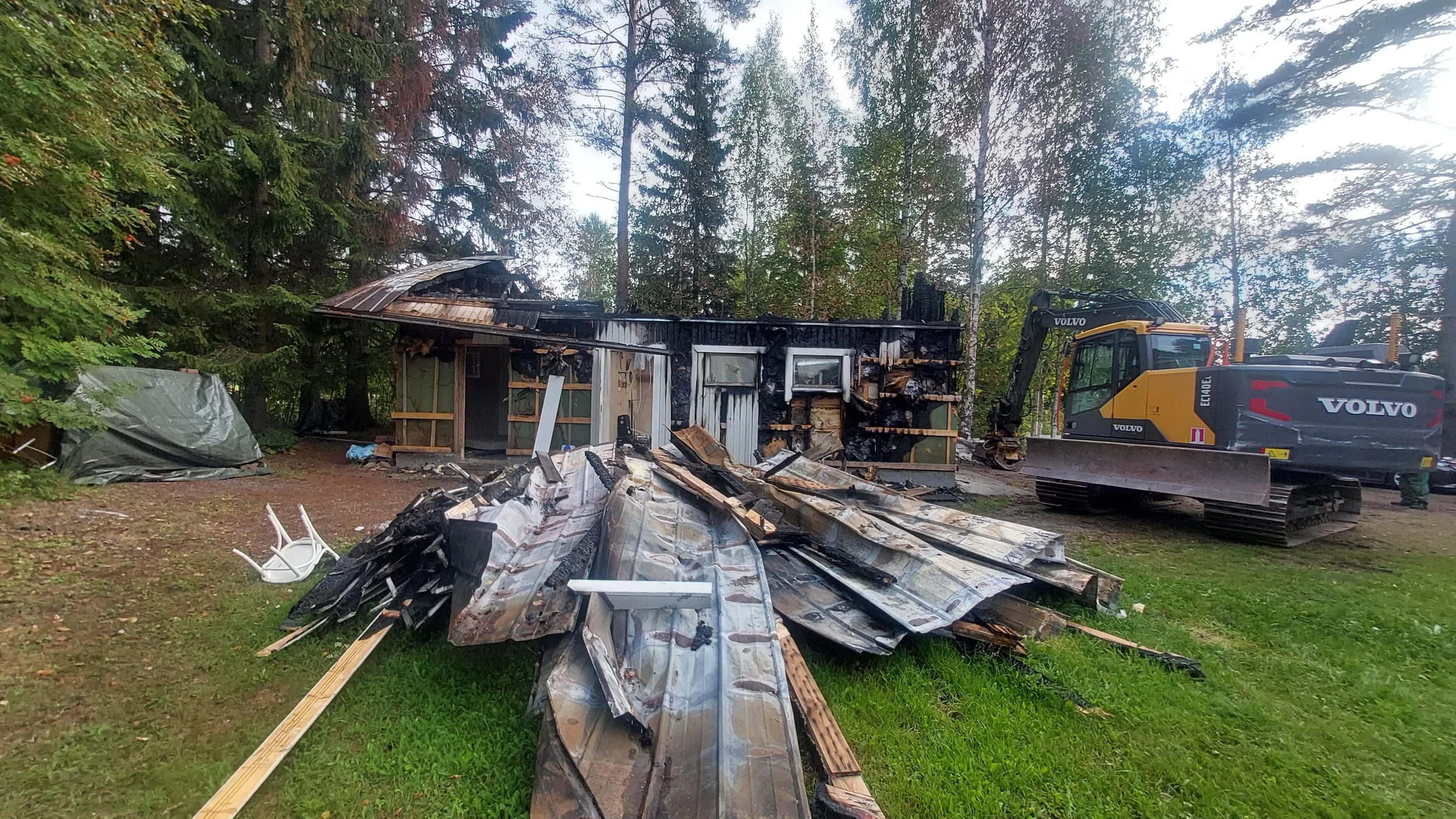 Takana kaivinkone ja sillä osittain purettu palanut talo, etualalla purkujätettä.
