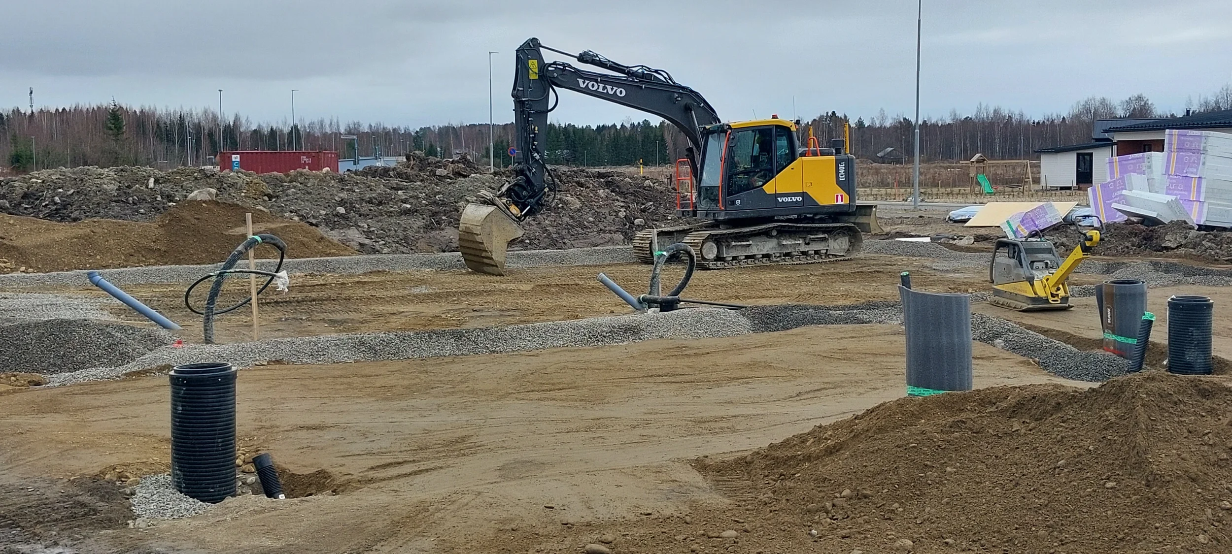 Kaivinkone tekee rakennuksen pohjatöitä rakennustyömaalla, ympärillä erilaisia rakennusmateriaaleja.