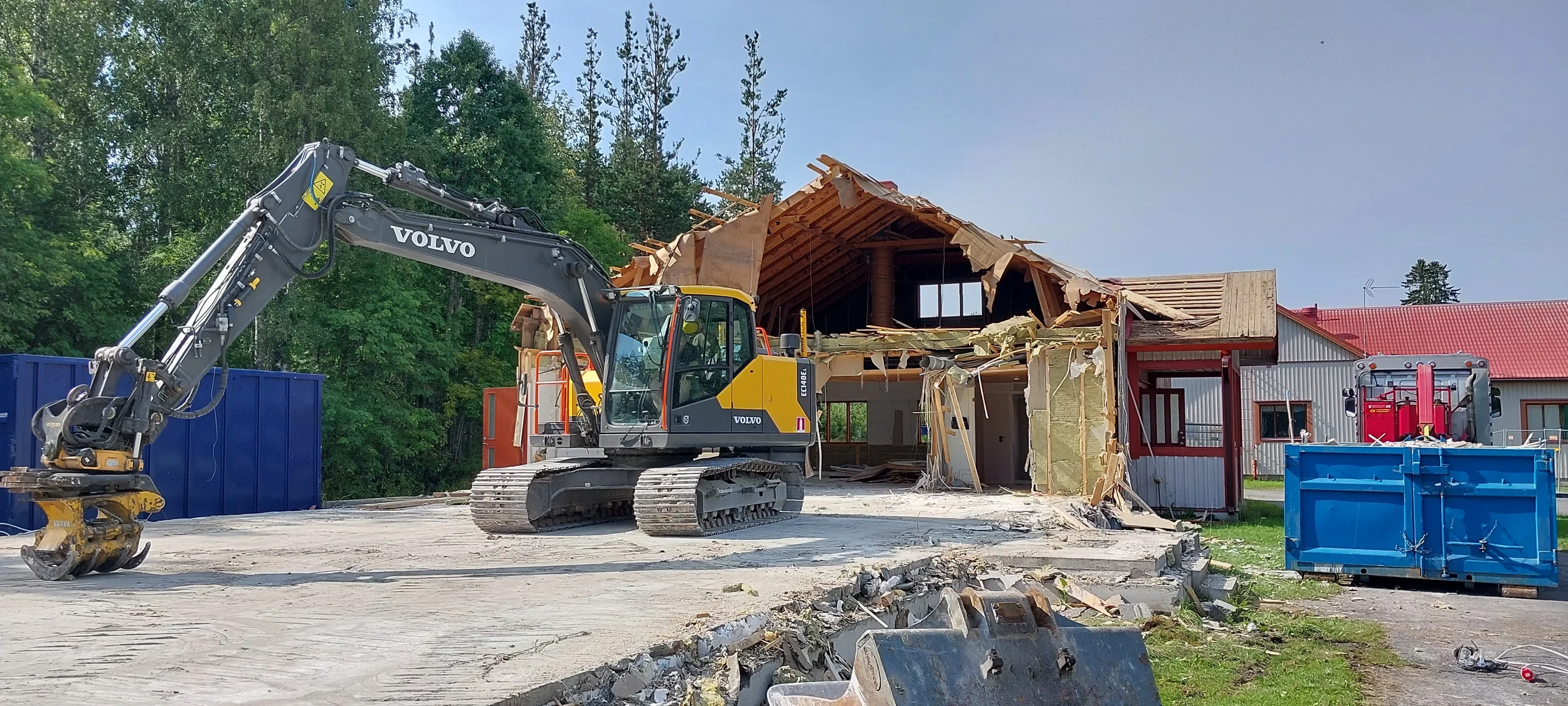 Purkutyömaa, jossa on kaivinkone, roskalavoja ja osittain purettu omakotitalo. Taustalla metsää ja omakotitalo, sininen taivas.