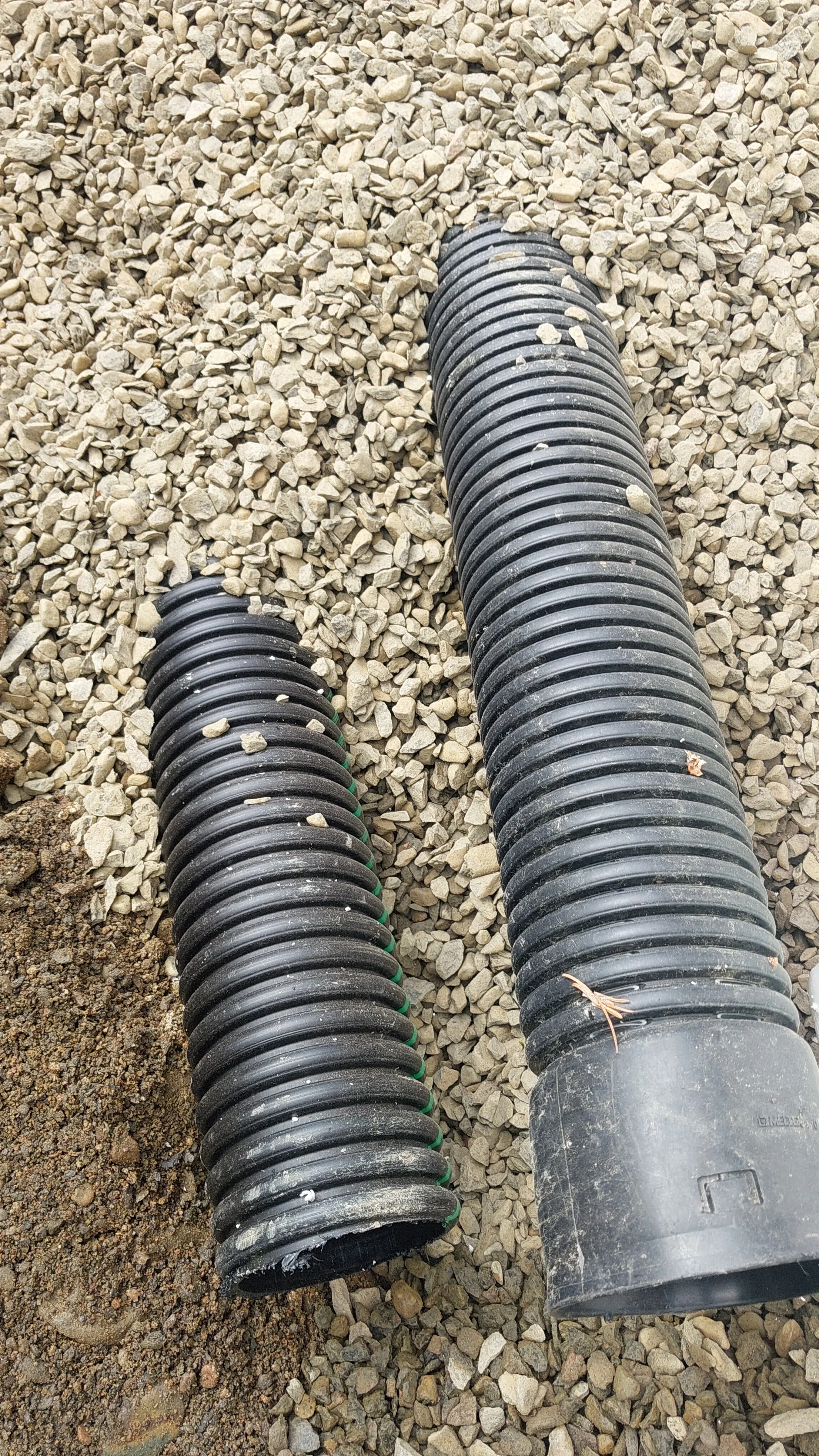 Kaksi mustaa muoviputkea salaojakaivannossa soran keskellä ja osittain soralla peitettyinä.