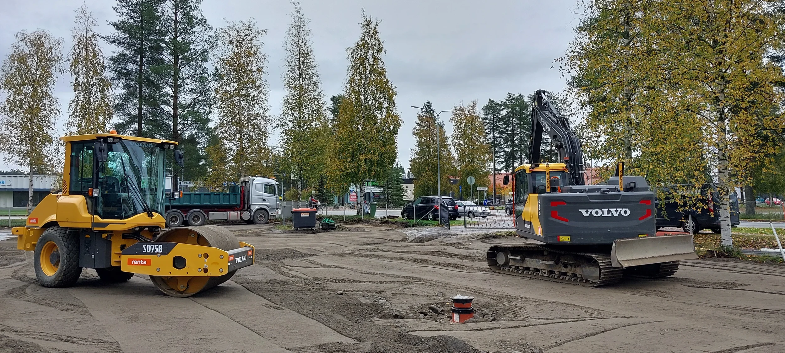 Jyrä ja Volvo merkkinen kaivinkone korjaamassa pihaa. Ympärillä puita ja liikennettä kaupungissa.