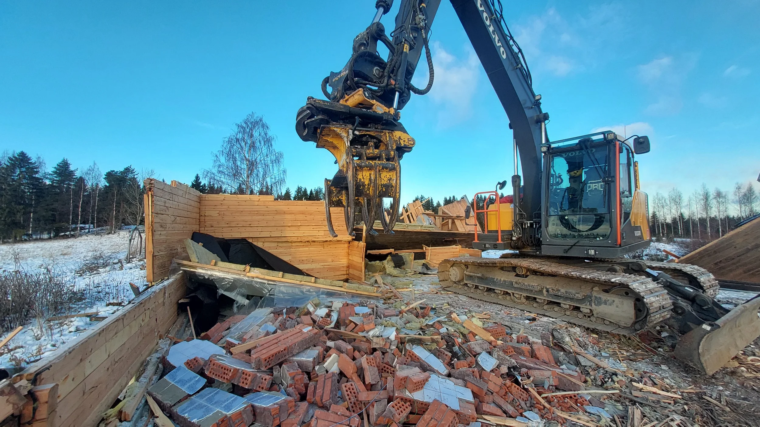 Kaivinkone purkaa purkutyömaalla rakennusta, maassa on tiili- ja puutavarajätettä. Taustalla kirkkaan sininen taivas.