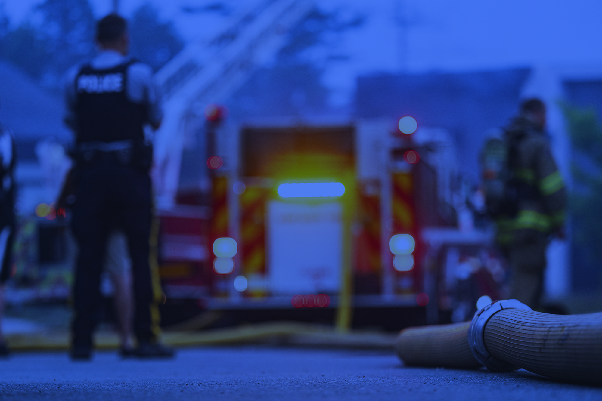 Blurred scene of police officers and emergency responders at night, with a fire truck and a rolled-up fire hose on the ground.