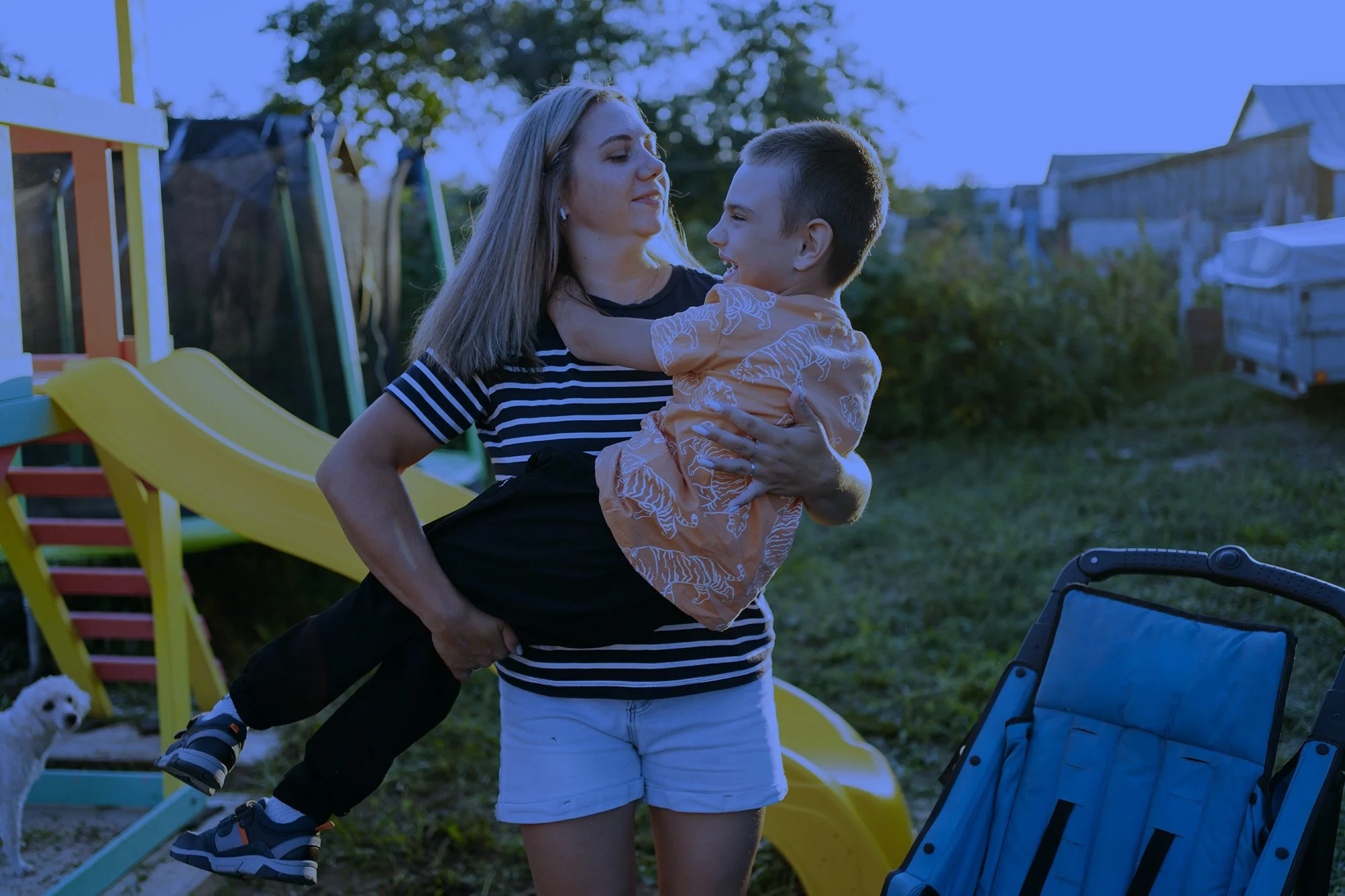 A woman holding a young boy in her arms outdoors near a playground. The woman is wearing a striped shirt, and the boy is wearing an orange shirt with animal prints. There is a yellow slide, a small dog, and trees in the background.