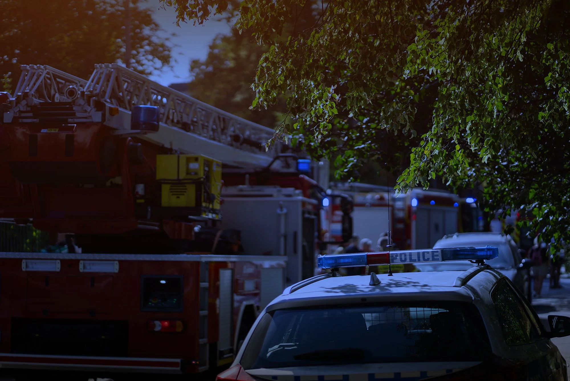 Photo of multiple emergency response vehicles, including fire trucks and police cars, parked outdoors under green leafy trees with a partly cloudy sky.