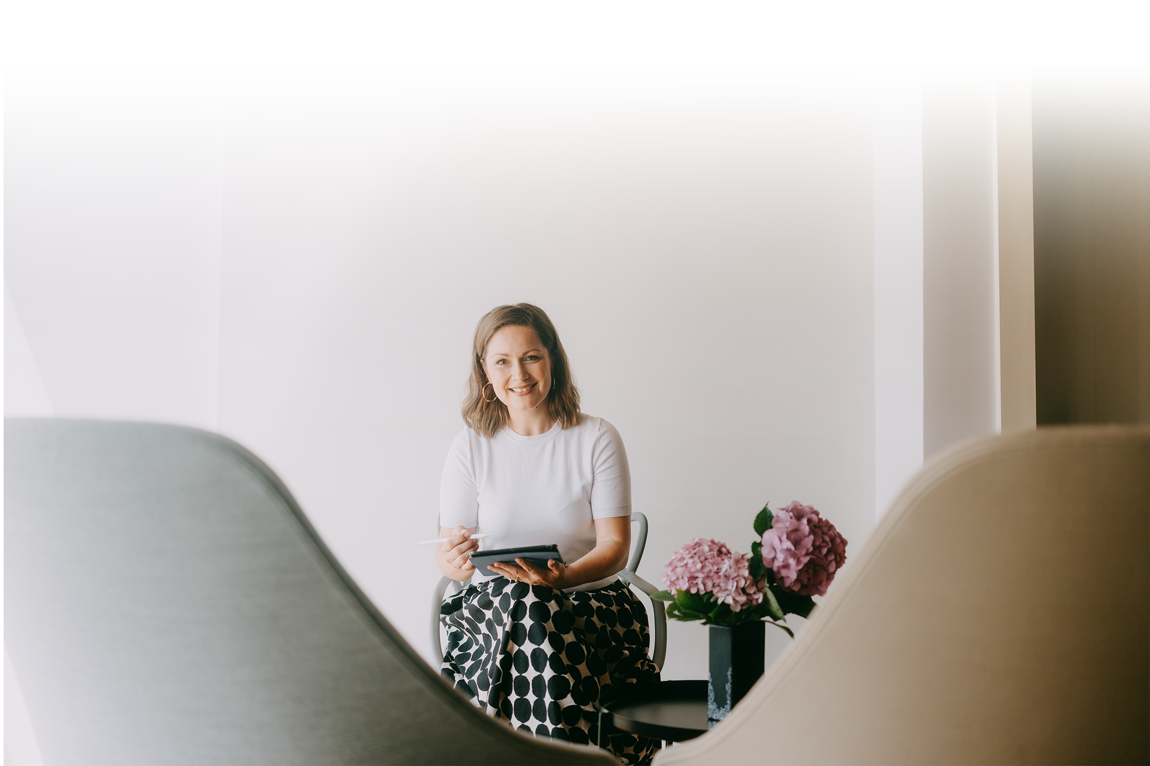 A woman with shoulder-length brown hair wearing a white top and polka dot skirt, holding a notepad and pen, sitting at a table with pink hydrangea flowers in the foreground, in a bright room with white walls.