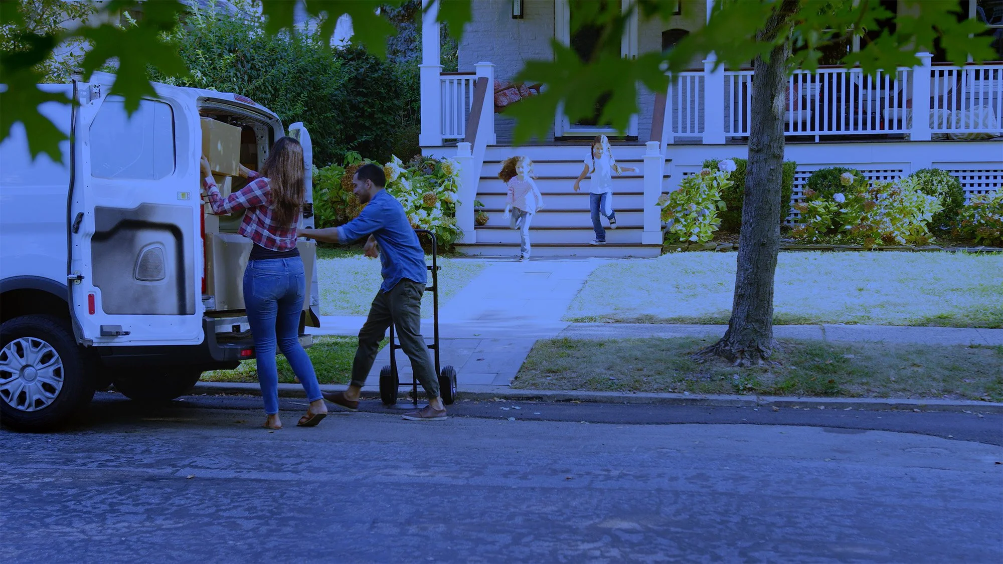 Family loading moving truck outside house, with two children walking down stairs, greenery around.