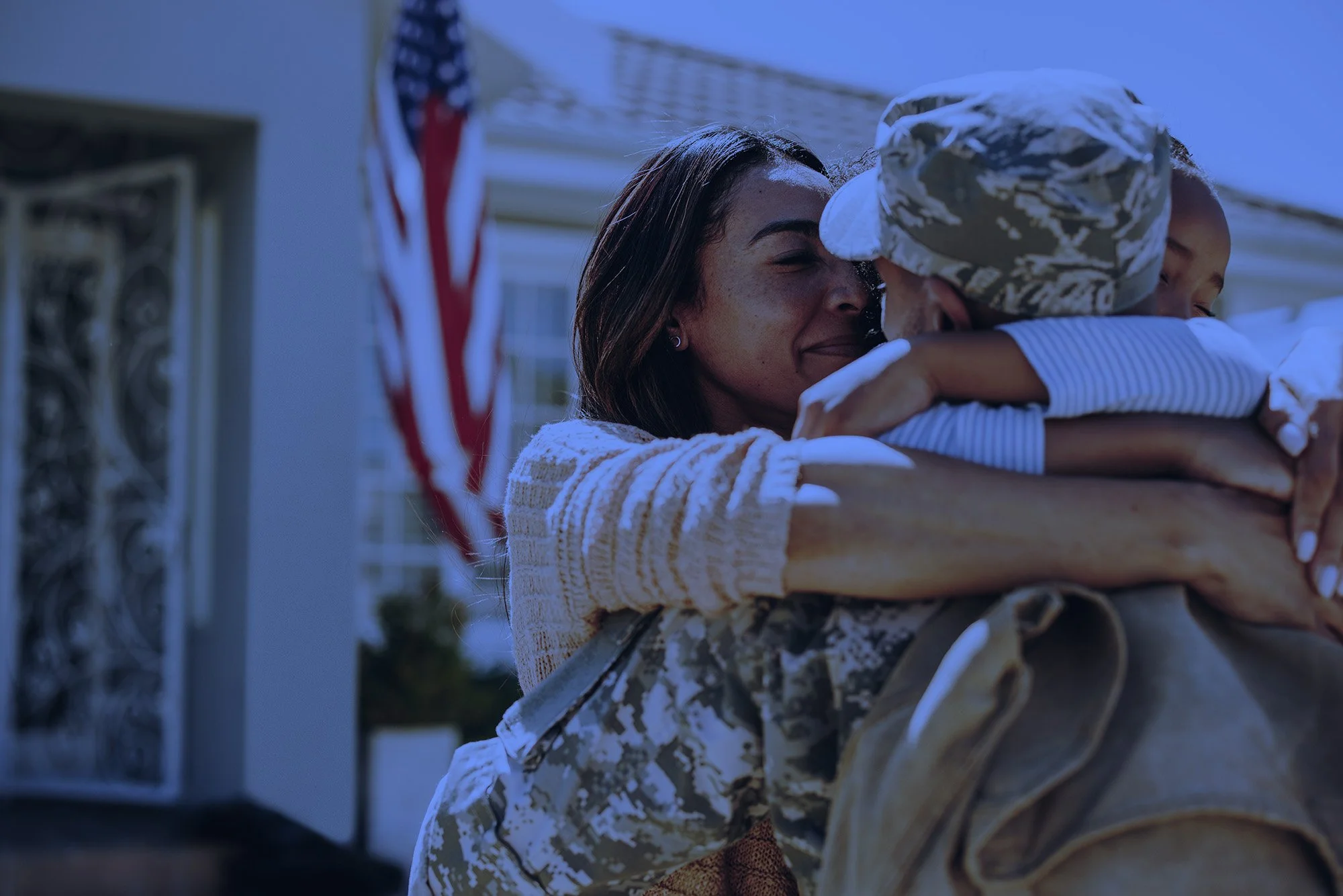 A woman in camouflage uniform embracing a young girl, possibly during a welcoming or burial ceremony. The girl is wearing a white and light blue striped shirt and a white cap, and is smiling with her eyes closed. An American flag is visible in the background.