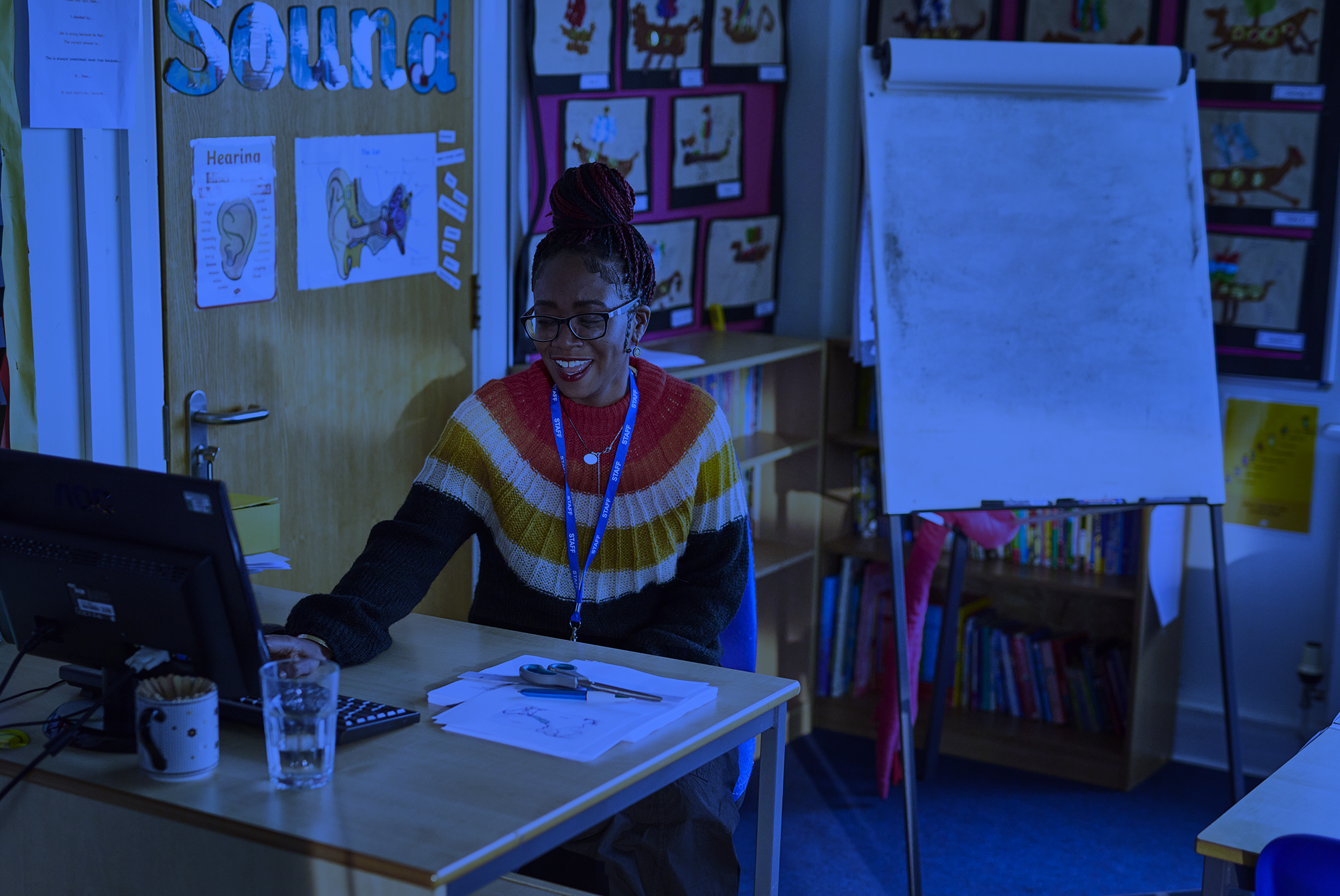 A woman with glasses and braided hair wearing a striped sweater, sitting at a desk in a classroom. She is smiling and working on papers. The background features educational posters, a whiteboard, and bookshelves.