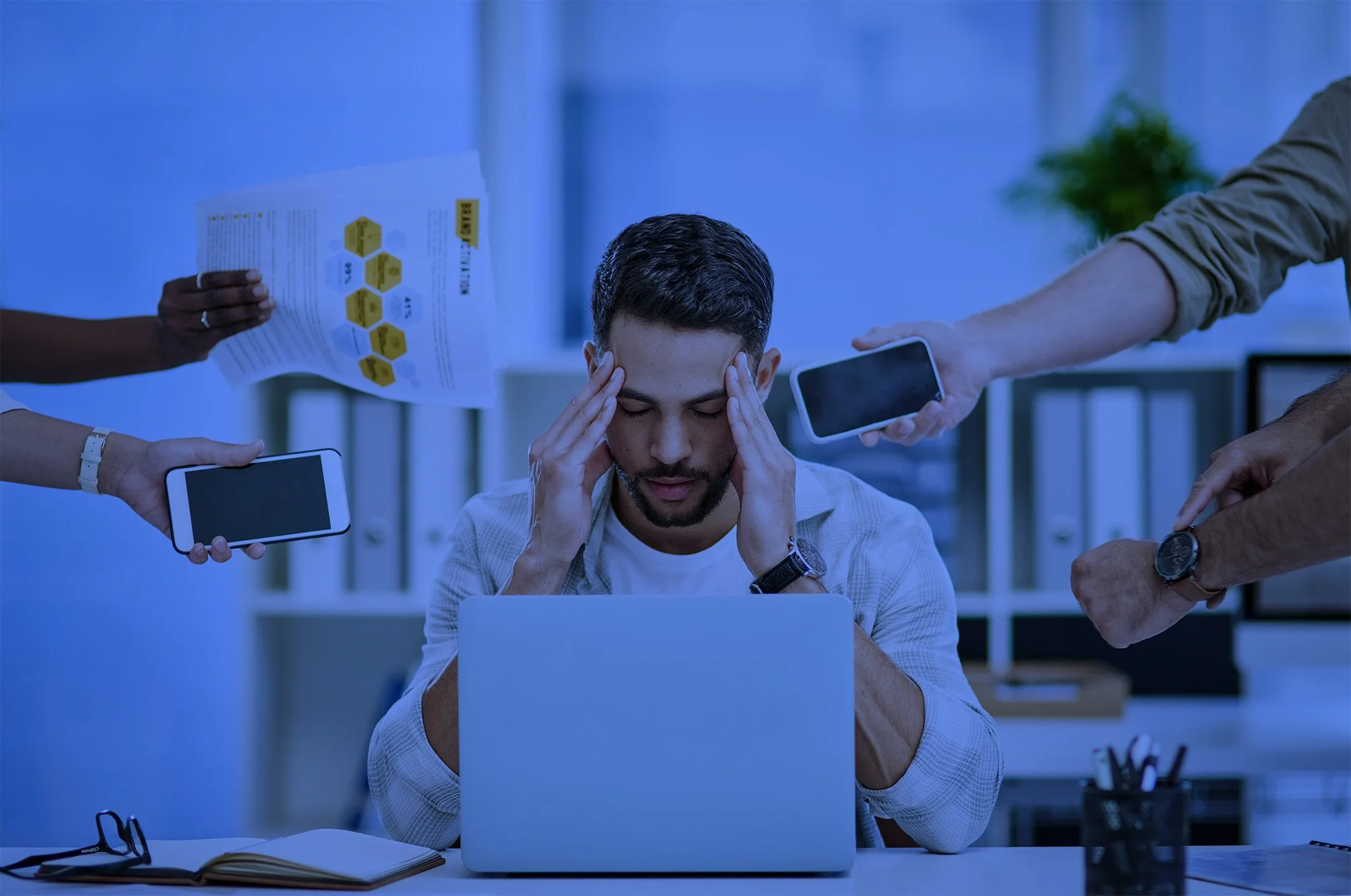 Man working at a laptop looks stressed as multiple hands reach out towards him with phones, papers, and a tablet in a busy office.