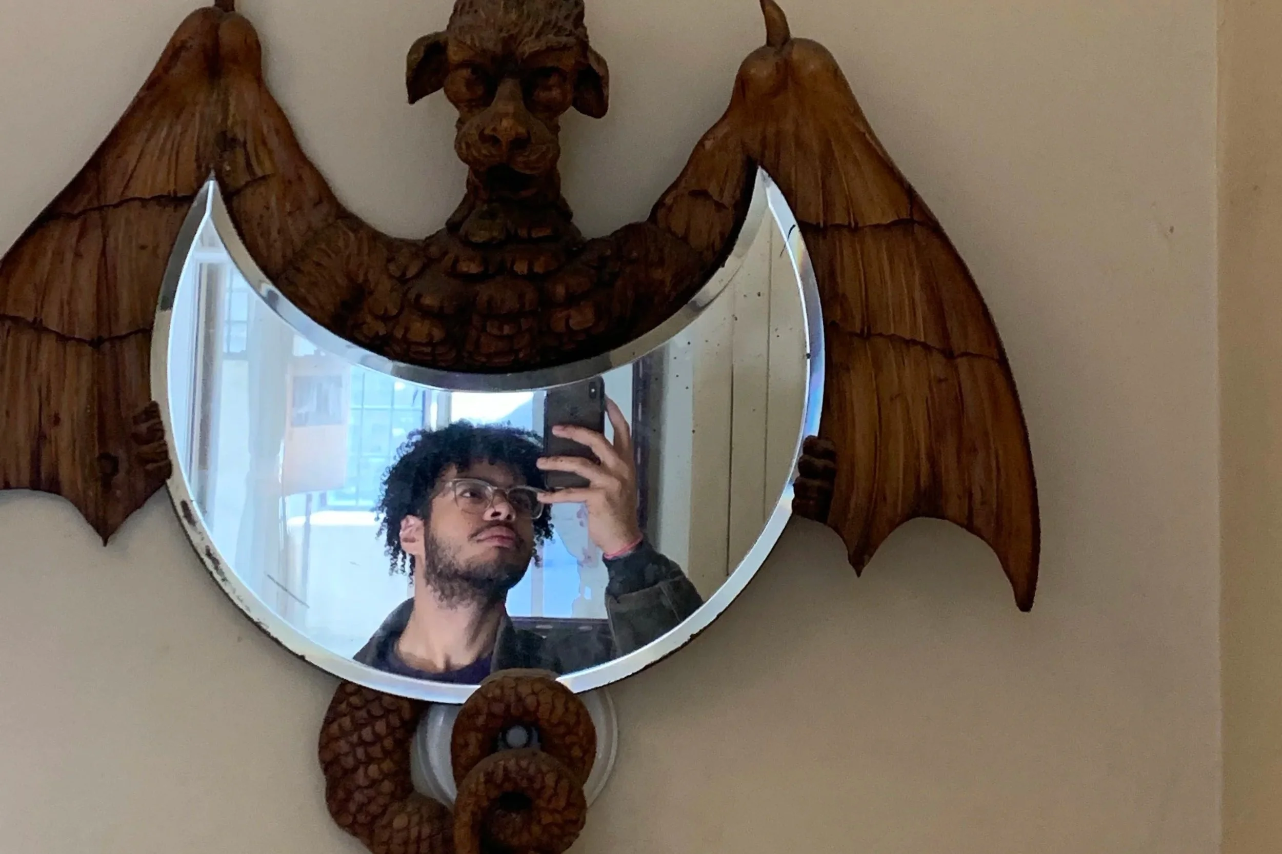 A round mirror with a wooden bat motif frame featuring a carved wolf's head. A man with curly dark hair, glasses, and a beard is taking a selfie in the mirror, with a background of a bright room and windows.