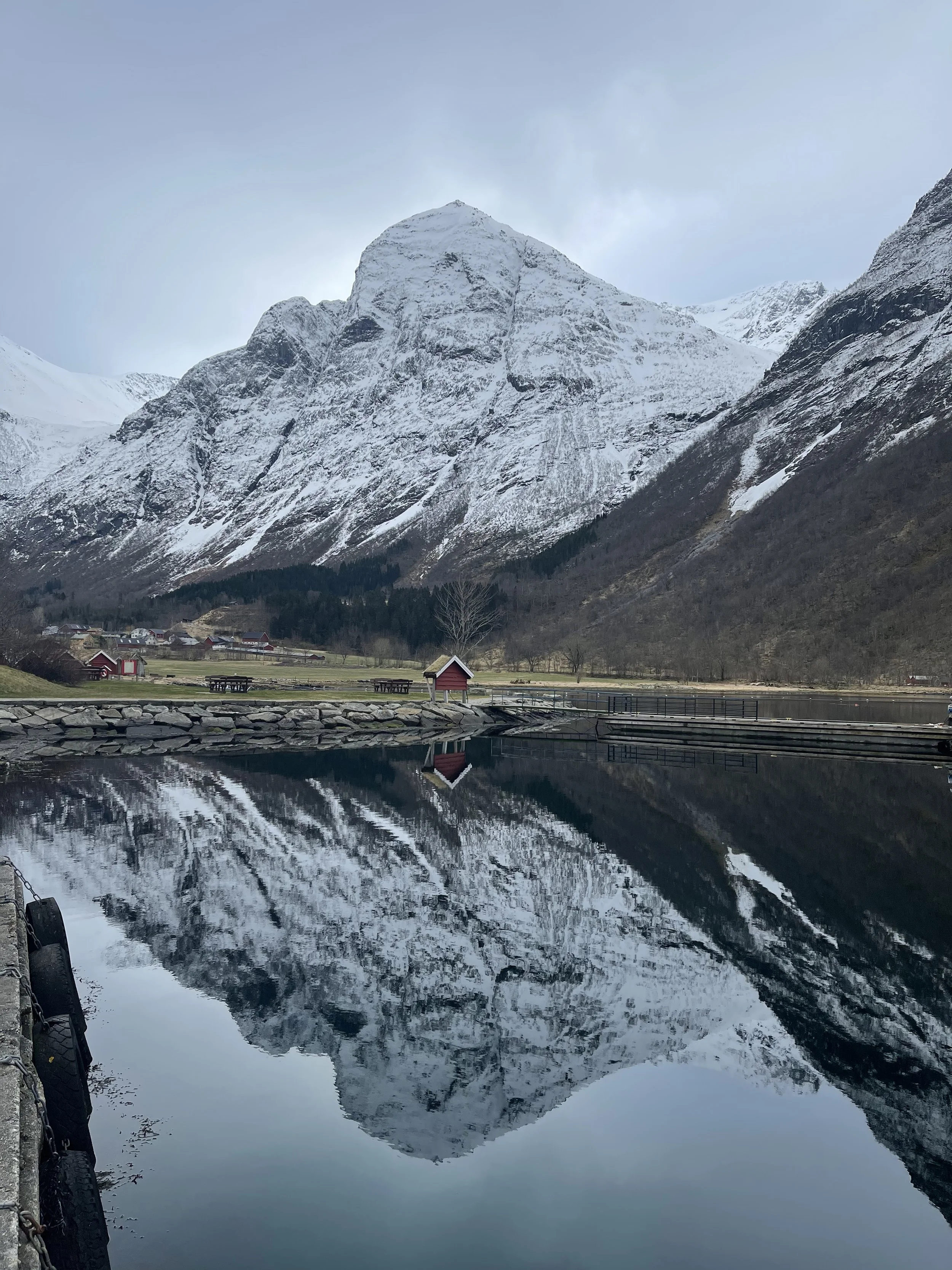 Snow-covered mountain reflecting in a calm body of water with a small red building and benches nearby.