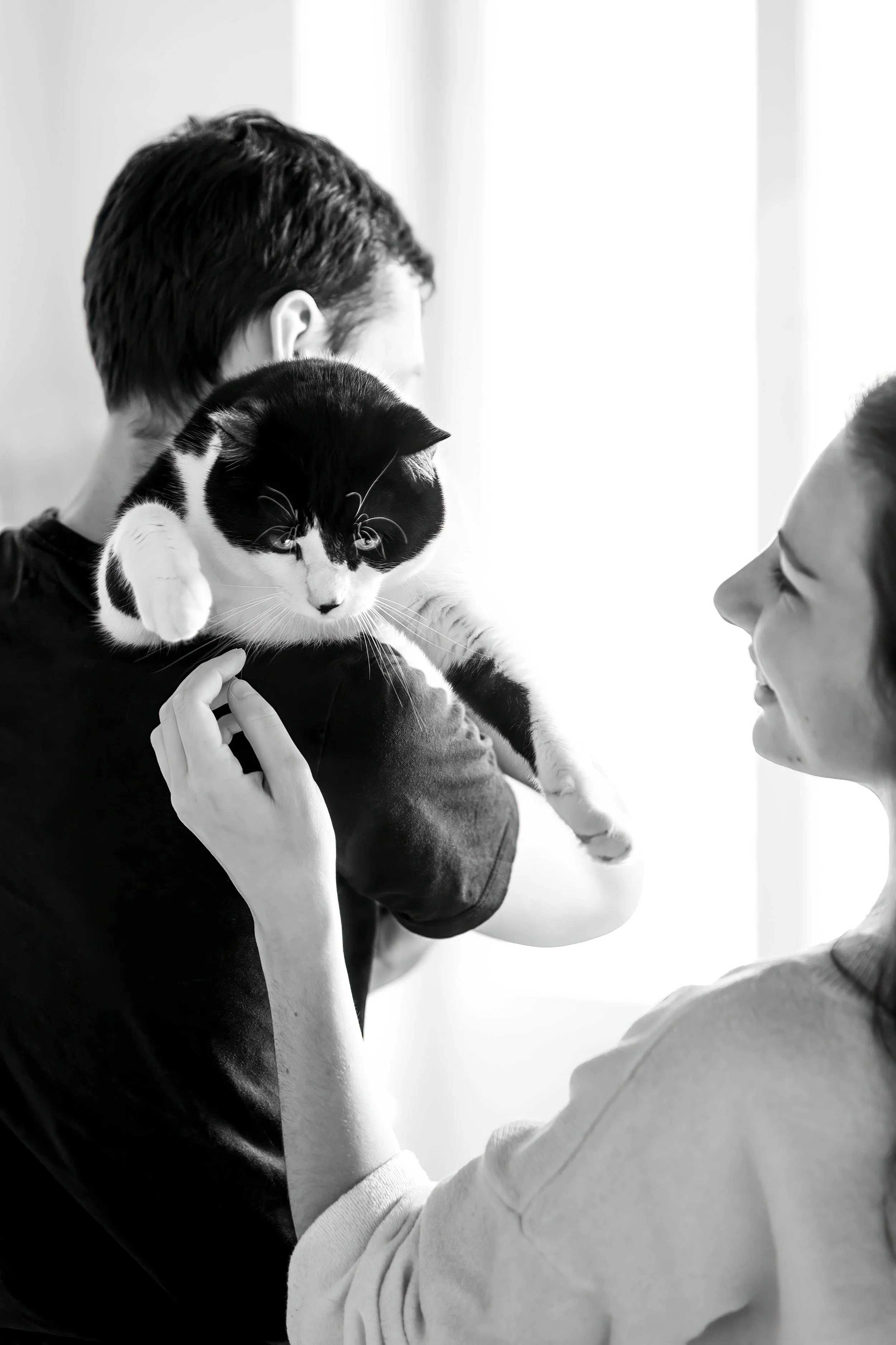 Un chat dans les bras d'un homme de dos et qui intéragit avec une femme souriante. Photo noir et blanc.