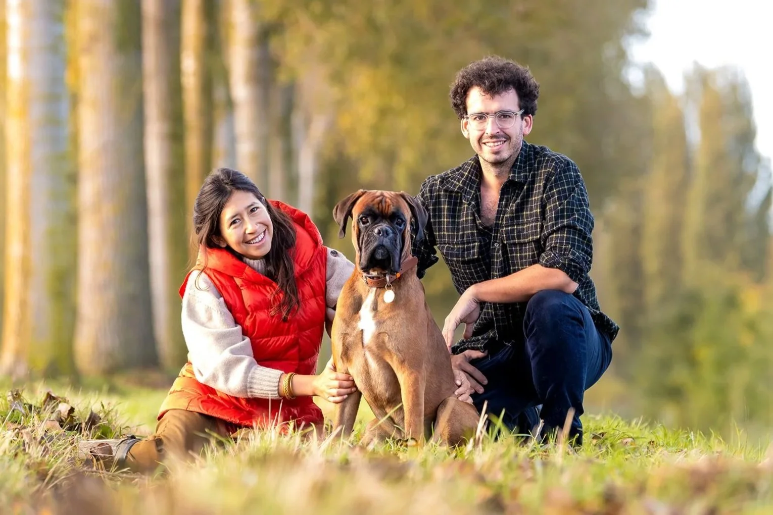 Un couple avec un chien boxer dans un parc en automne, souriant et posant pour une photo. 