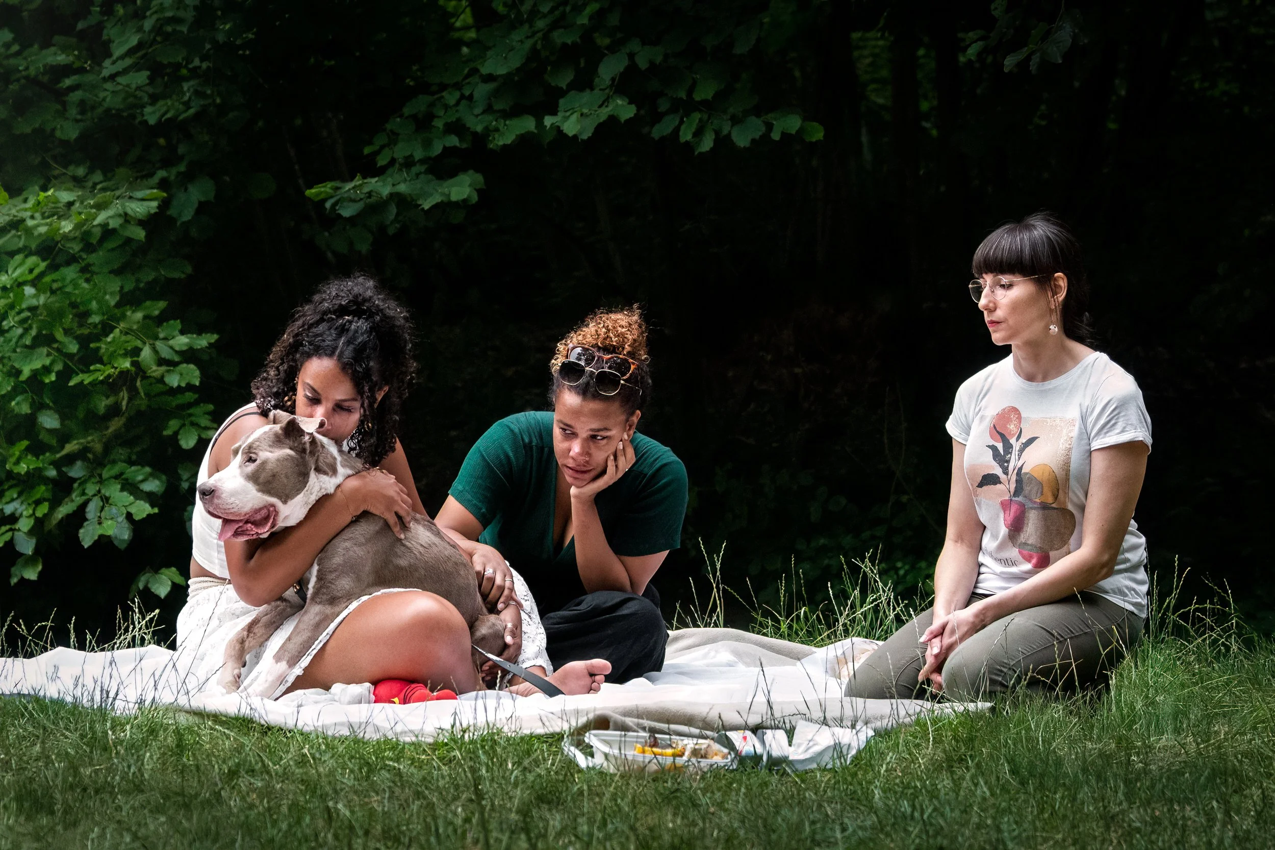 Trois femmes assises sur une couverture en plein air, deux d'entre elles caressent un chien de race staff qui est sur leurs jambes, un fond de forêt dense.