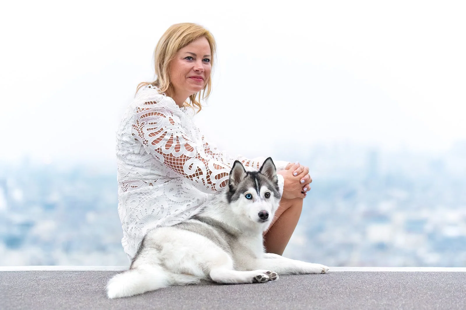 Une femme en robe blanche assise dans la rue à côté d'un chien pomsky allongé, avec une ville floue en arrière-plan. 