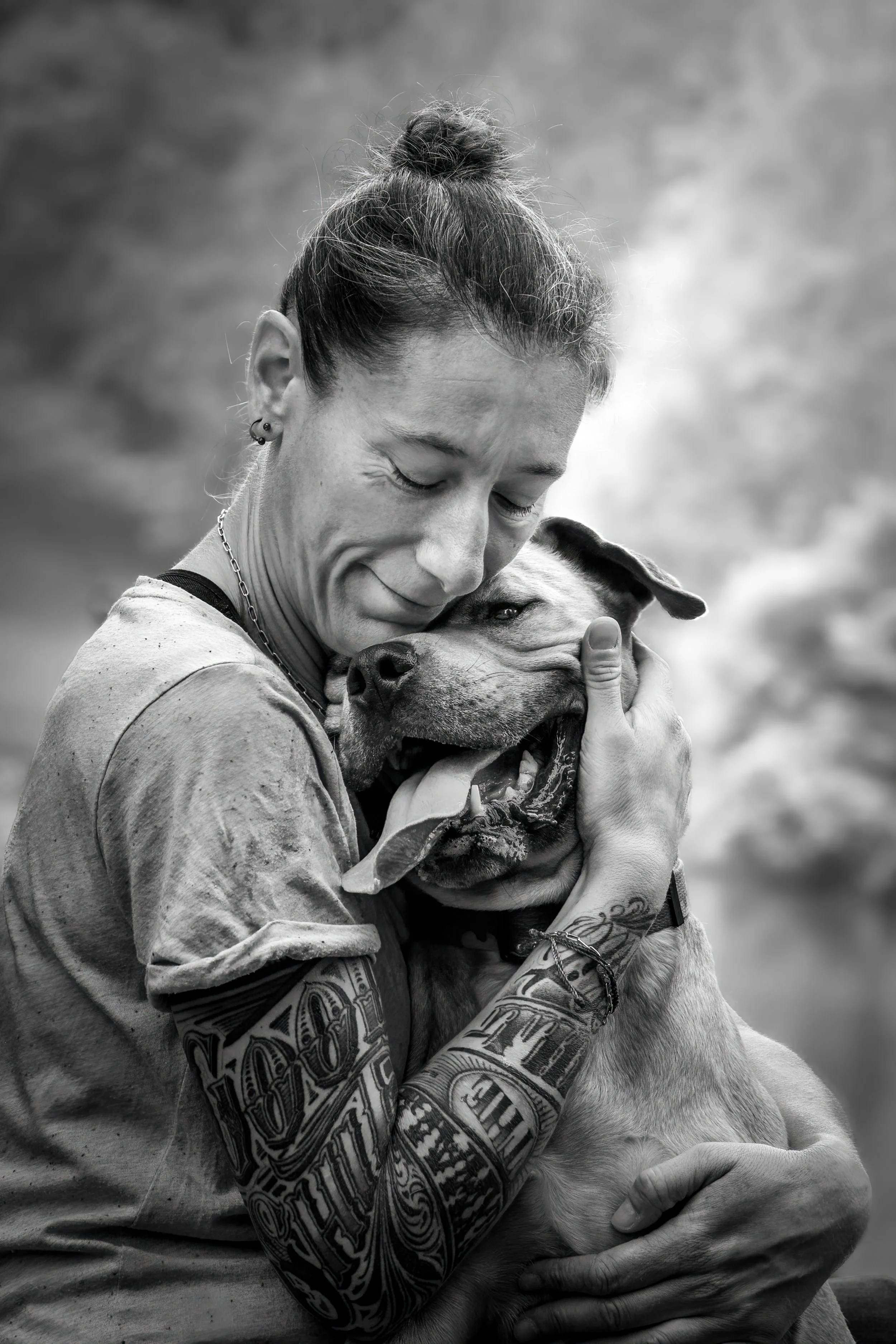 Une femme avec des tatouages embrasse affectueusement un chien staff, tous deux souriants, en noir et blanc. 