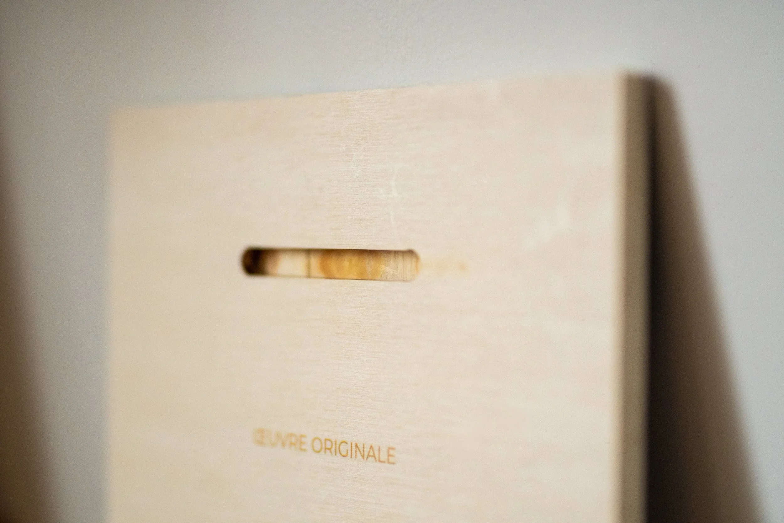 Le dos d'un tableau en bois clair avec une fente horizontale pour accrocher le tableau, et au centre et le mot « œuvre originale » écrit en dessous.