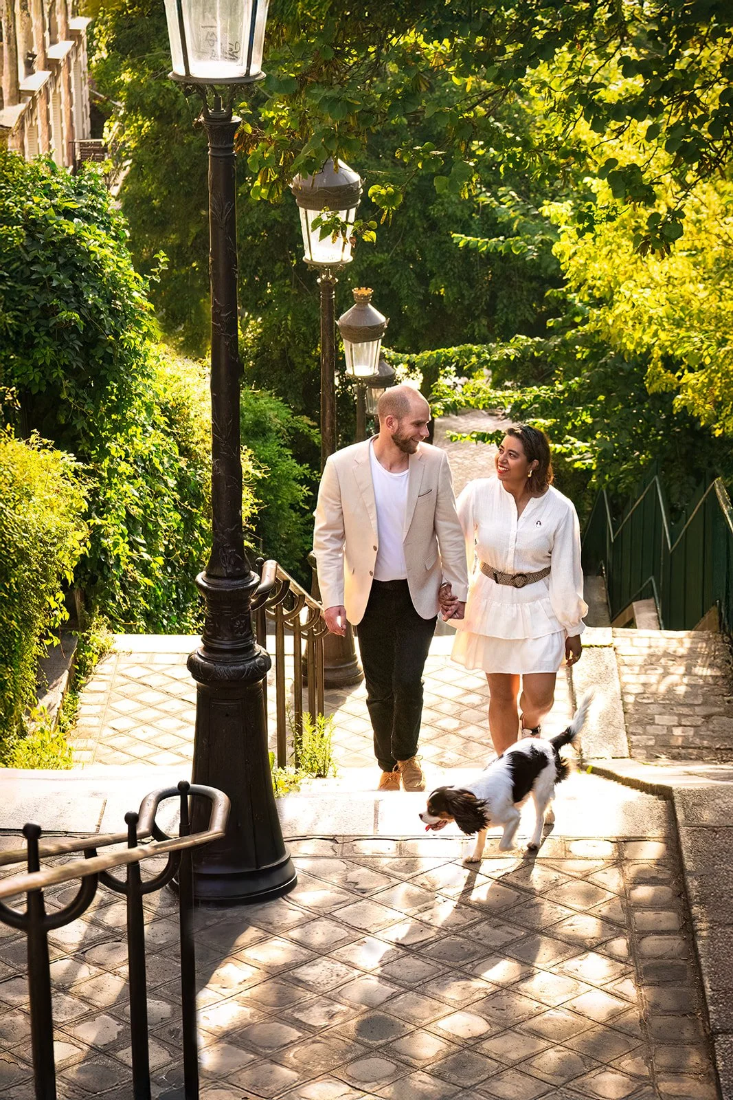 Un couple marchant dans un escalier de rue ensoleillé en se tenant la main, accompagné d'un chien cavalier king charles noir et blanc tenu en laisse, sous des lampadaires et entourés de verdure en été. 