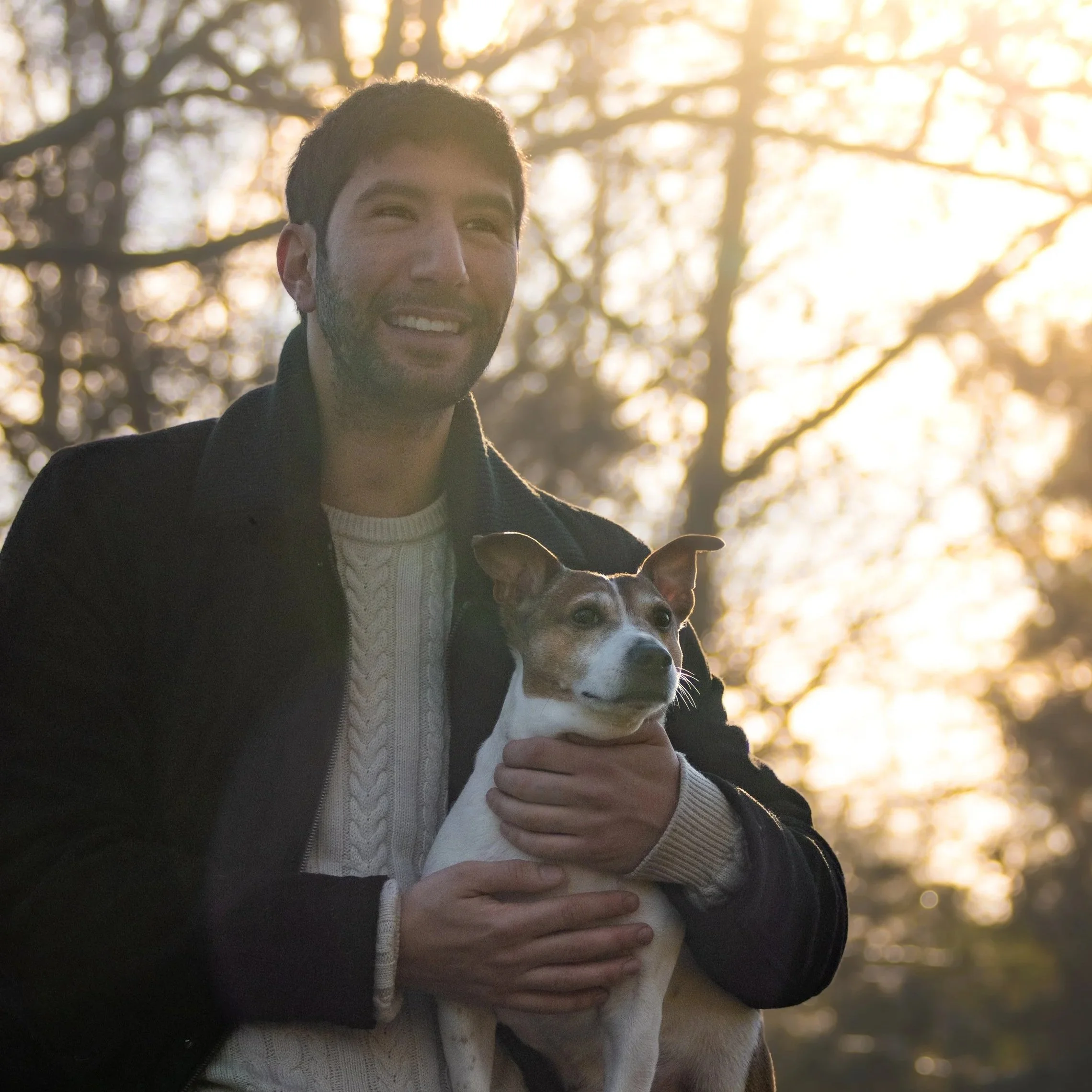 Un homme souriant tenant un chien jack russel, avec un arbre en arrière-plan et la lumière du soleil couchant. 