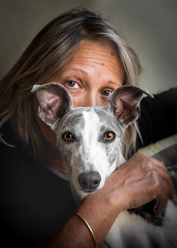 Une personne âgée avec des cheveux gris tient un chien whippet près de son visage, et regarde l'objectif, dans un intérieur sobre. 