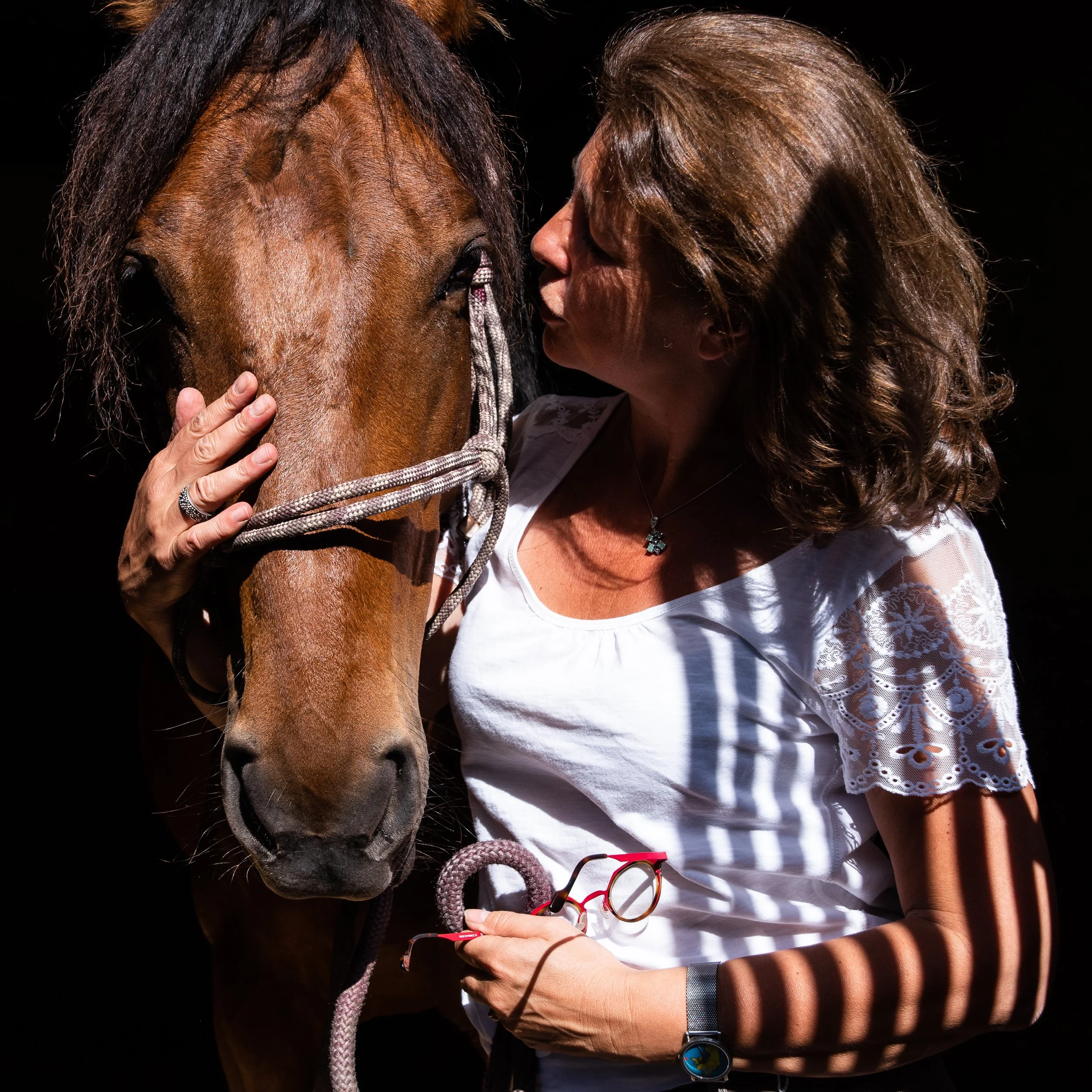 Une femme embrasse un cheval bai, en tenant sa tête, dans une lumière douce avec des ombres de rayures. 