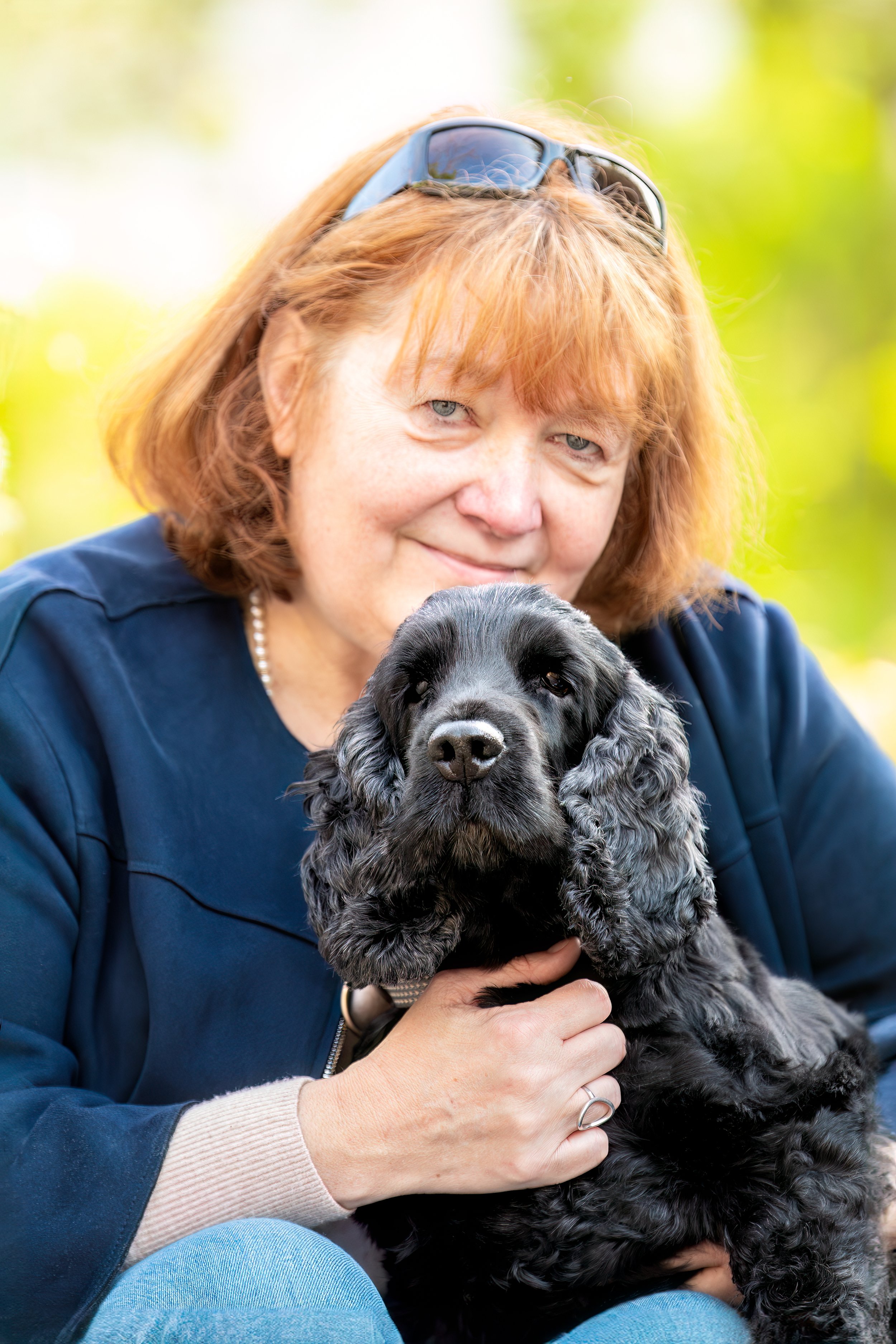 Une femme tient un chien cocker noir à oreilles longues dans ses mains, en extérieur, avec un fond flou de verdure. 