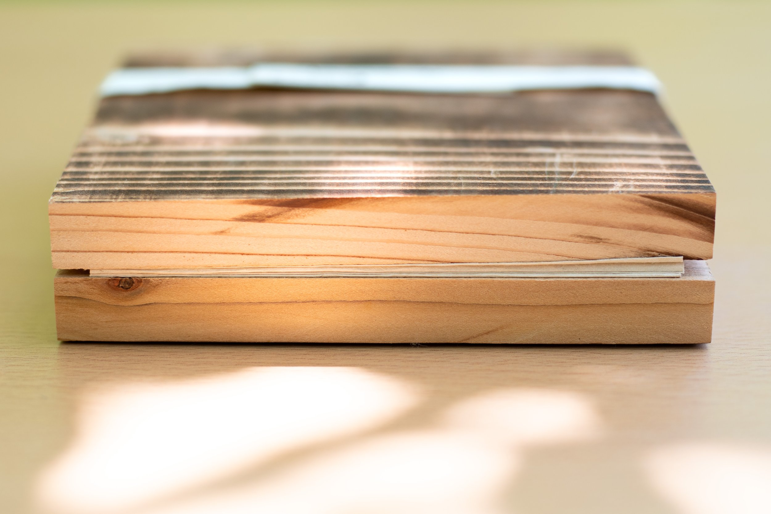 Un livre leporello fermé avec les deux couvertures en bois, avec la surface supérieure en bois brûlé, posée sur une surface en bois clair. La lumière naturelle crée des reflets et des ombres douces autour de l'objet.
