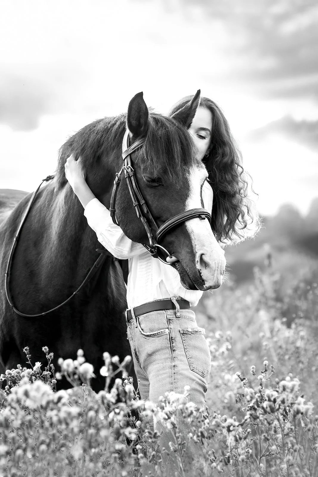 Une jeune femme embrasse et étreint un cheval, qui a les yeux fermés, dans un champ avec un ciel nuageux. Photo noir et blanc.