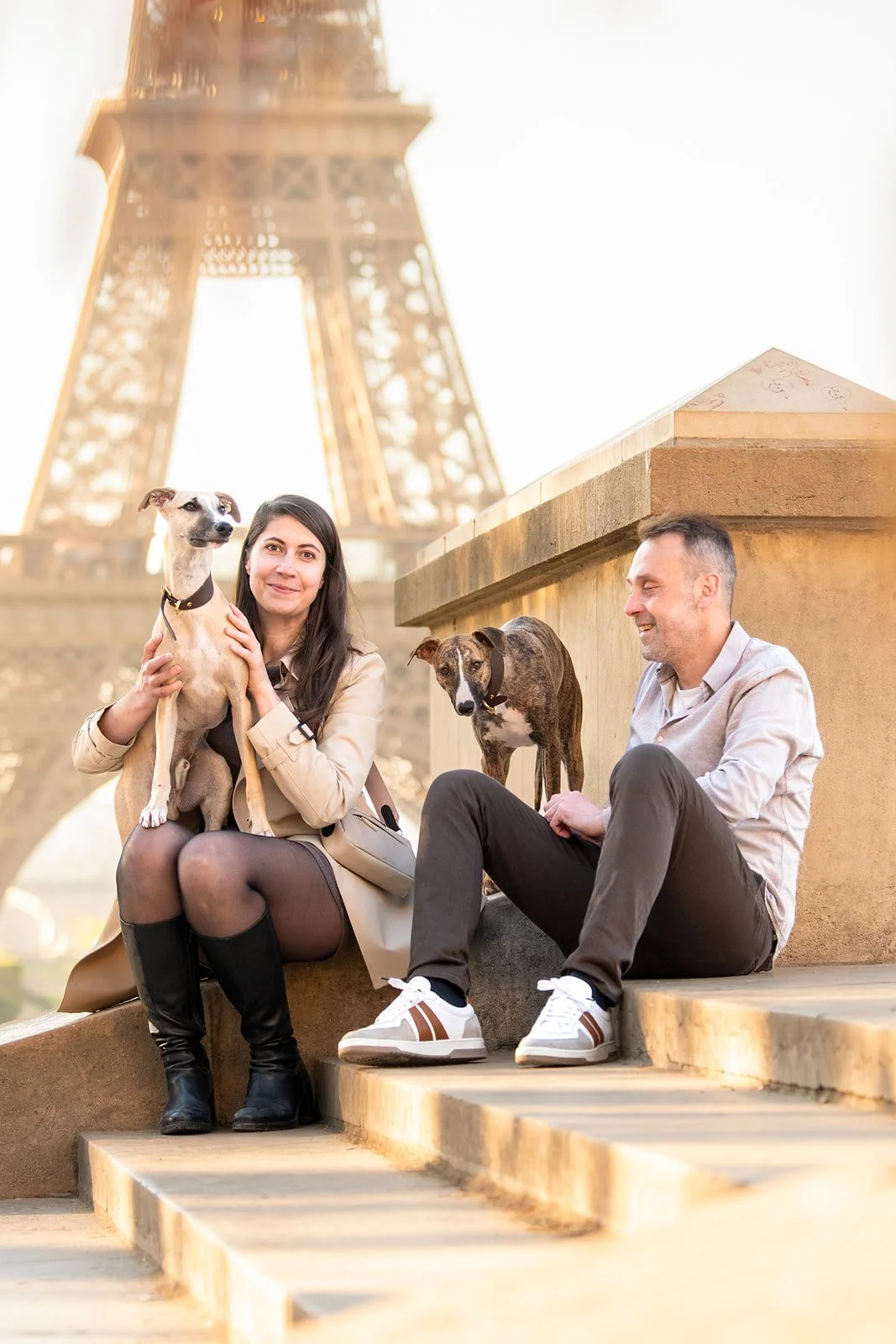 Un couple et deux chiens assis sur des marches près de la Tour Eiffel. La femme tient un whippet beige et blanc, l'homme tient un whippet bringé. 
