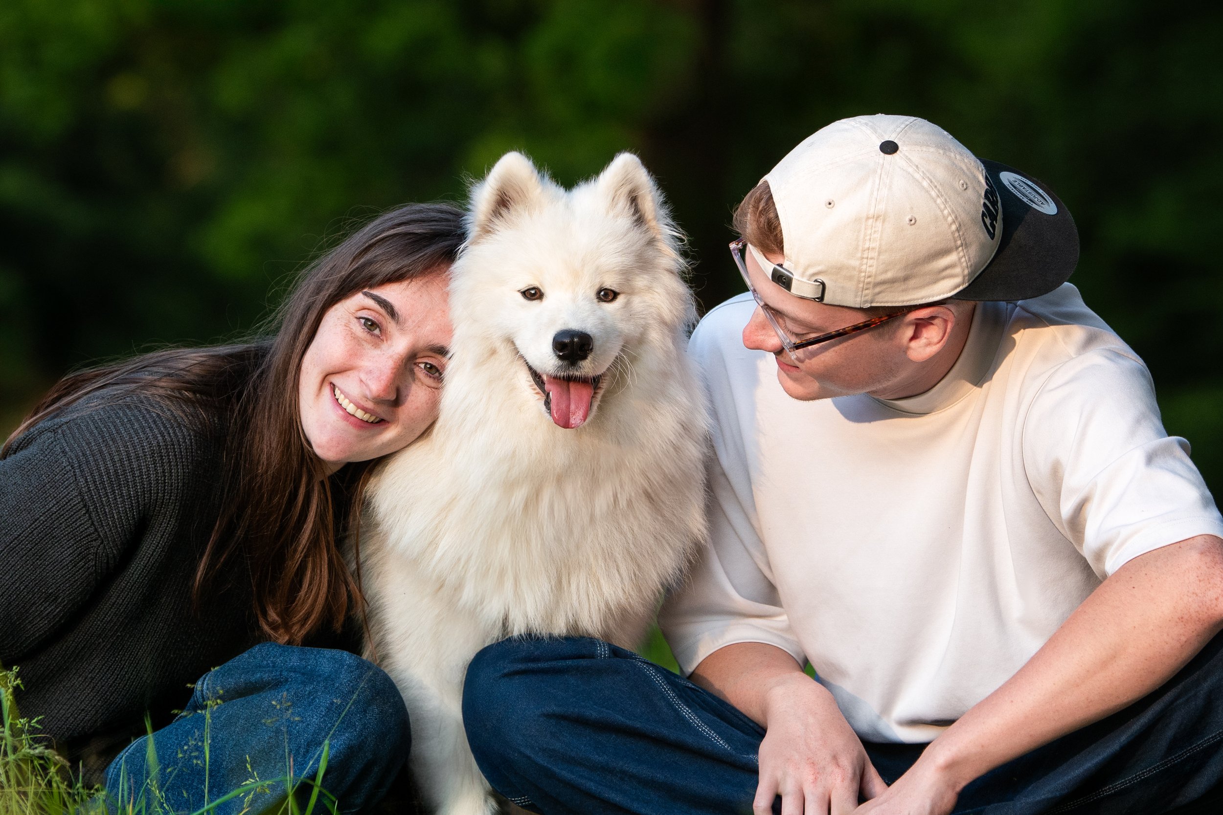 Une jeune femme, un jeune homme avec casquette et un chien samoyède blanc assis dans l'herbe, souriant et profitant d'une sortie en plein air.