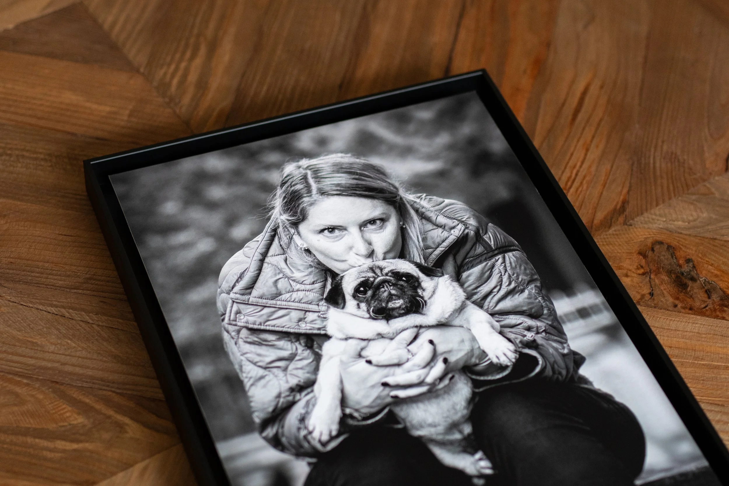 Cadre d'une photographie en noir et blanc d'une femme tenant et embrassant un petit chien carlin, regardant l'objectif, posée sur un fond flou