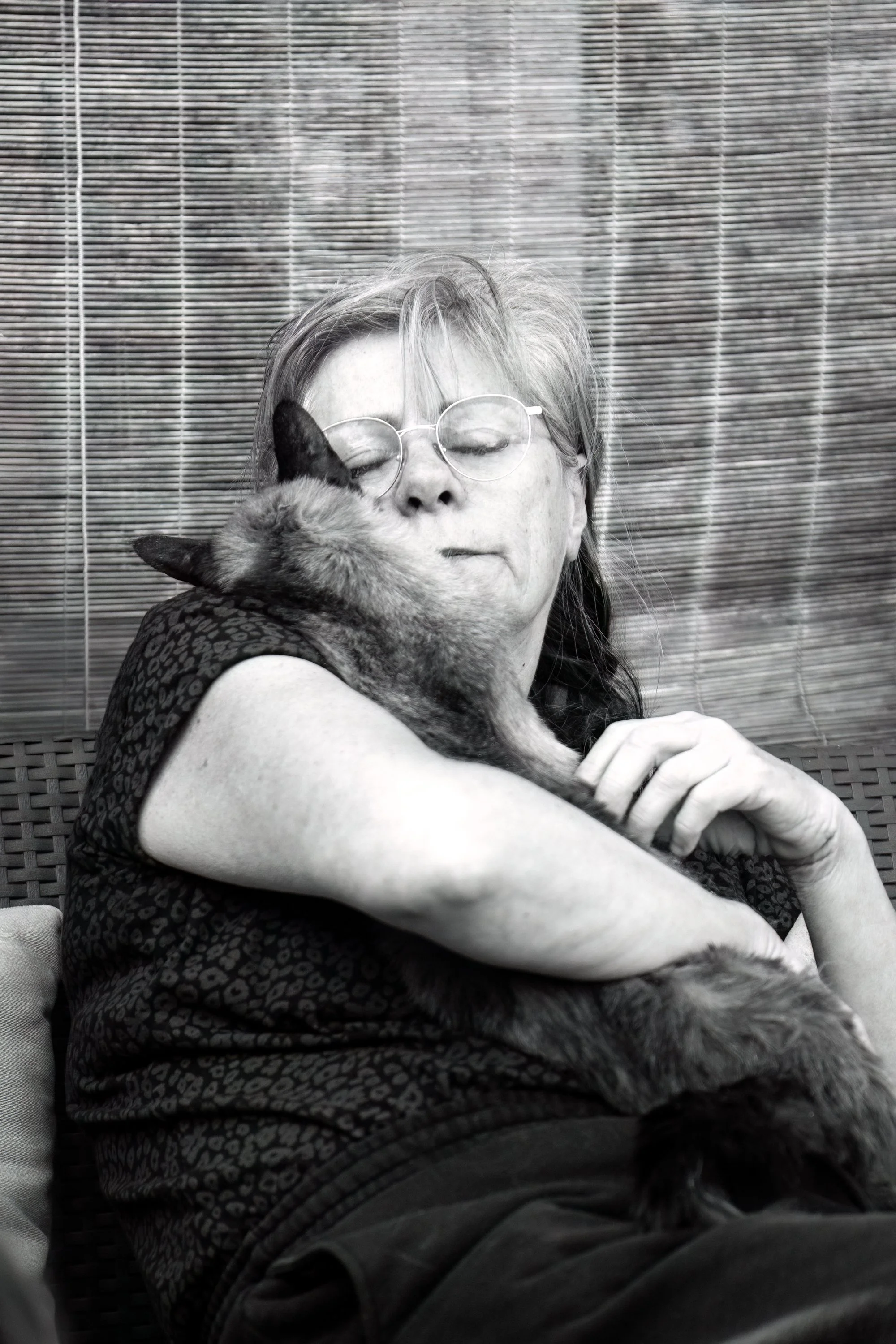 Une femme avec des lunettes tient un chat dans ses bras et le serre tendrement, dans une cour extérieure. La photo est en noir et blanc.