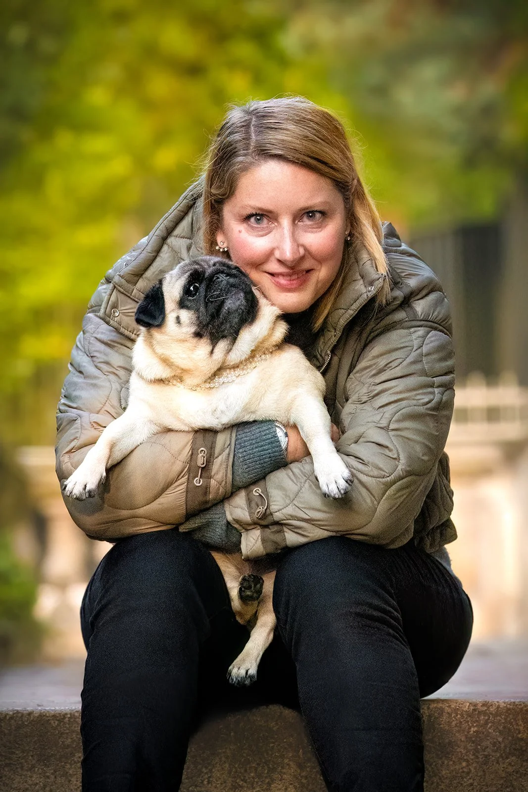 Une femme souriante assise sur des marches tient un chien carlin beige sable et noir dans ses bras, en extérieur avec un fond flou de feuillage vert. 