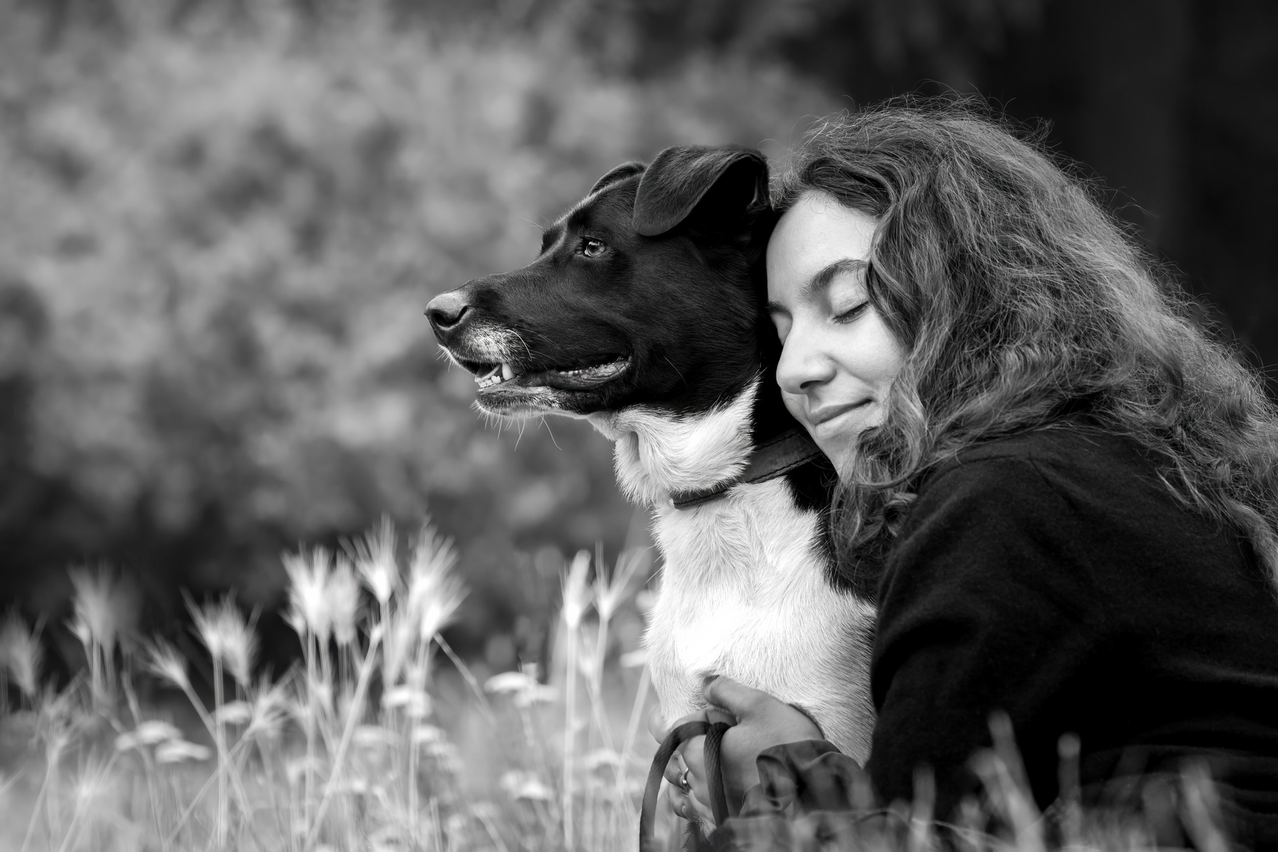 Une femme et un chien assis dans un champ d'herbes, partageant un moment de complicité avec la femme qui a les yeux fermés et sourit. Photo noir et blanc.