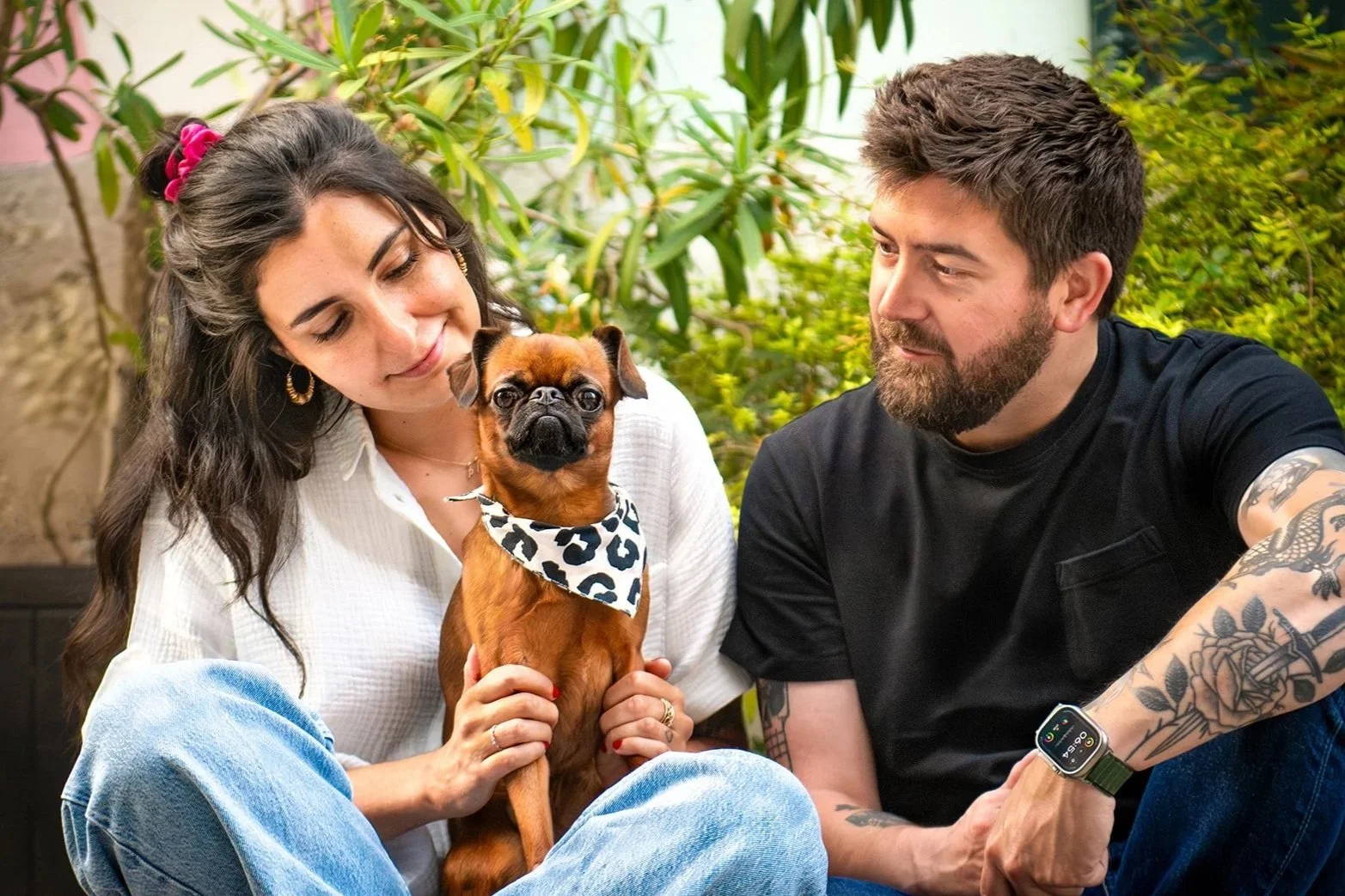 Un couple assis dans un jardin, la femme tient un chien marron et noir petit brabançon avec un bandana à motifs, et les deux humains regardent le chien avec affection. 