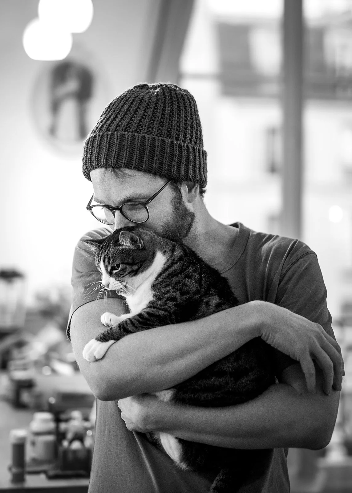 Un homme portant un bonnet en laine et des lunettes tient un chat dans ses bras, dans un décor intérieur. Photo noir et blanc.