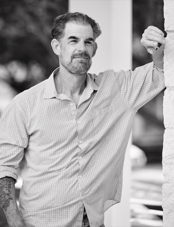 A black-and-white photo of a man with a beard and mustache, wearing a checked shirt, standing outdoors next to a wall, with trees and parked cars in the background.
