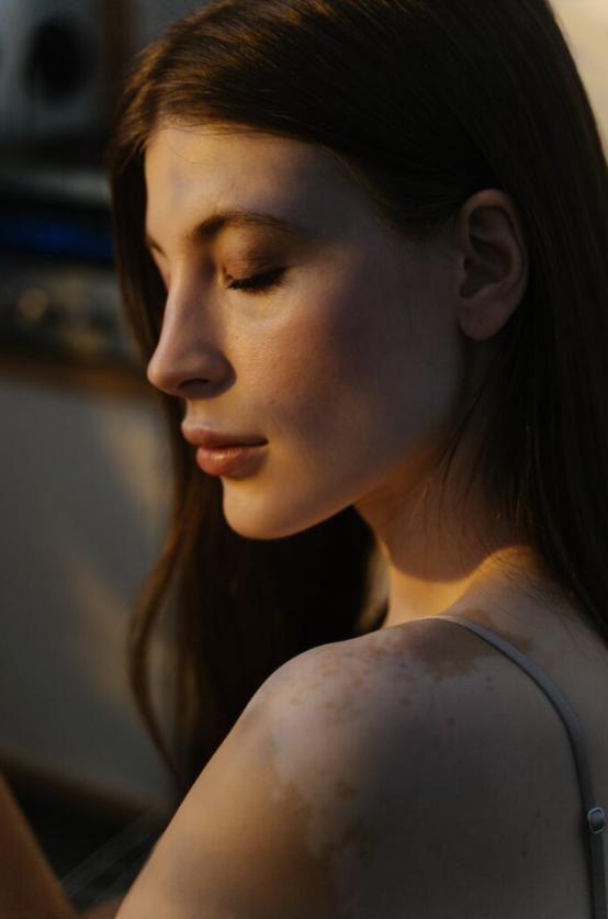 Close-up of a young woman with closed eyes, smiling slightly, with sunlight on her face and shoulder, in a soft, warm setting.