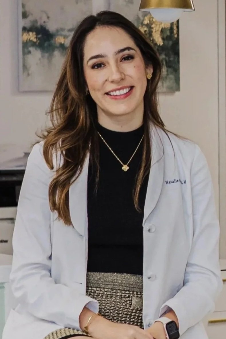 A professional portrait of Dr. Hone smiling. She has long, layered light brown hair and is wearing a white medical lab coat over a black high-neck top. She is accessorized with a delicate gold clover-shaped pendant necklace.
