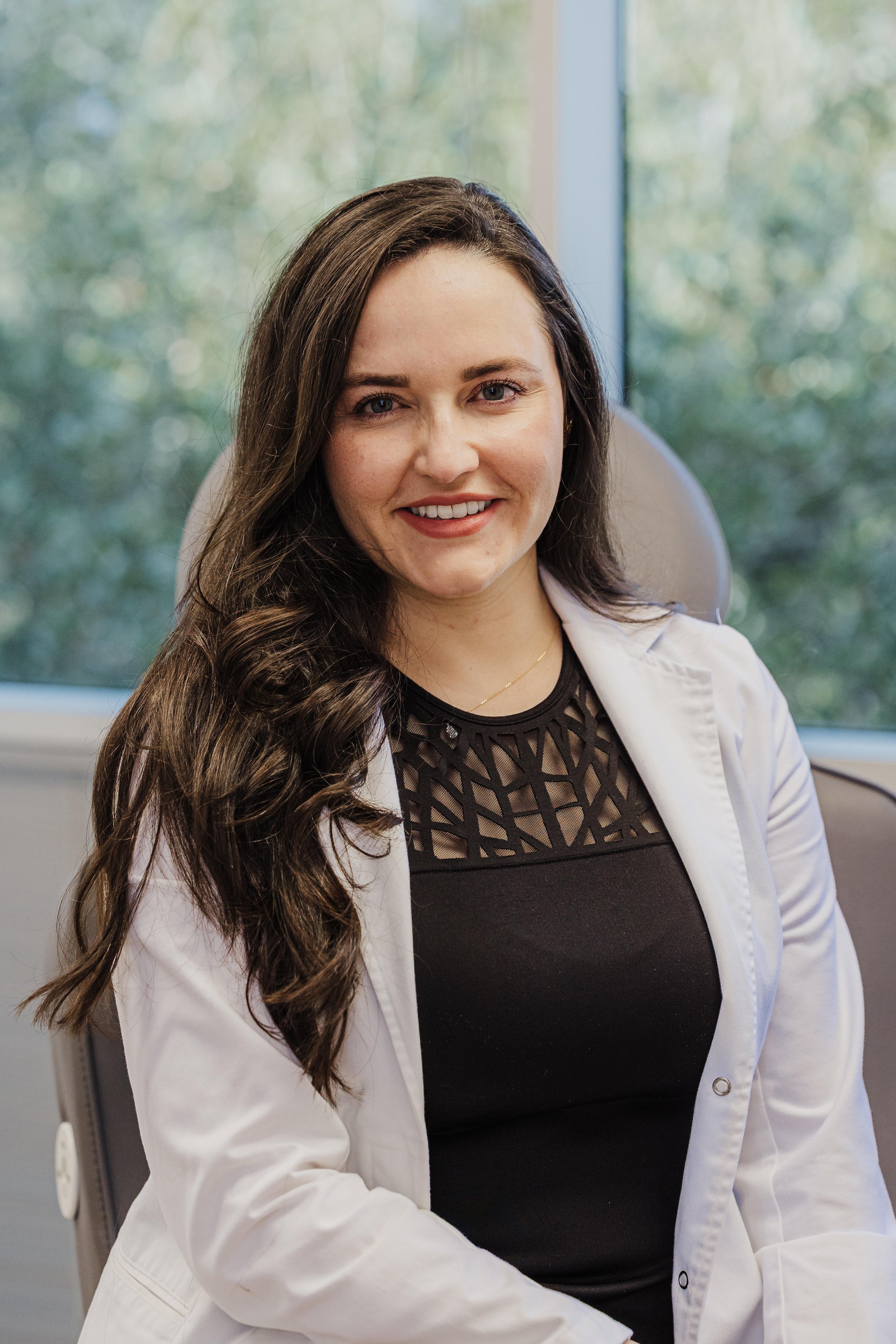 A professional portrait of Dr. Anita, a woman with long, wavy brown hair, smiling warmly. She is wearing a white medical lab coat over a black top with a geometric cutout neckline.