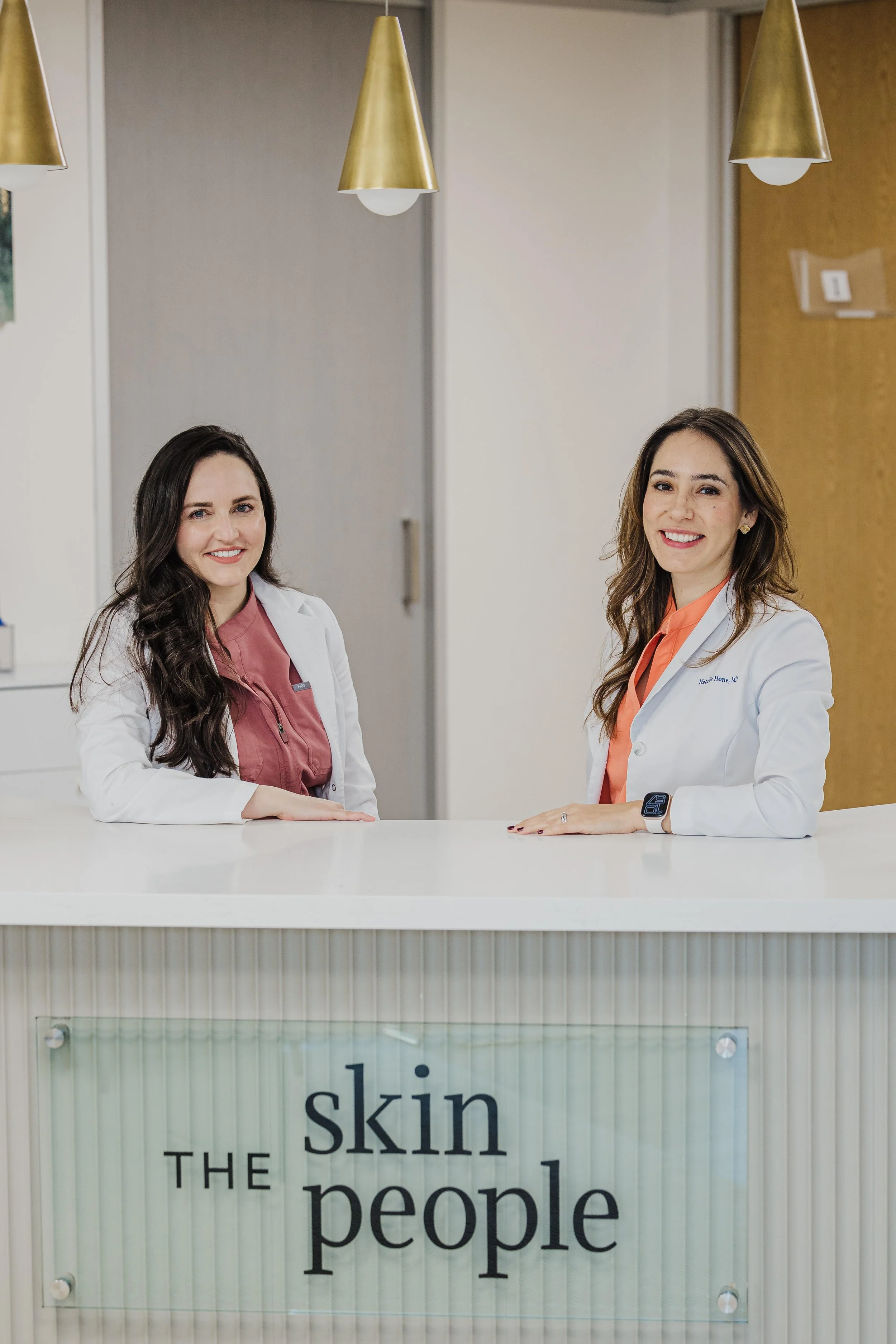 Two women in white lab coats standing behind a reception desk with a sign that reads "The Skin People". They are smiling and looking at the camera.