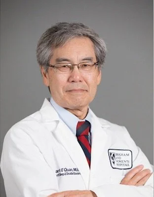 Portrait of a male doctor in a white coat with glasses, standing with arms crossed against a gray background.